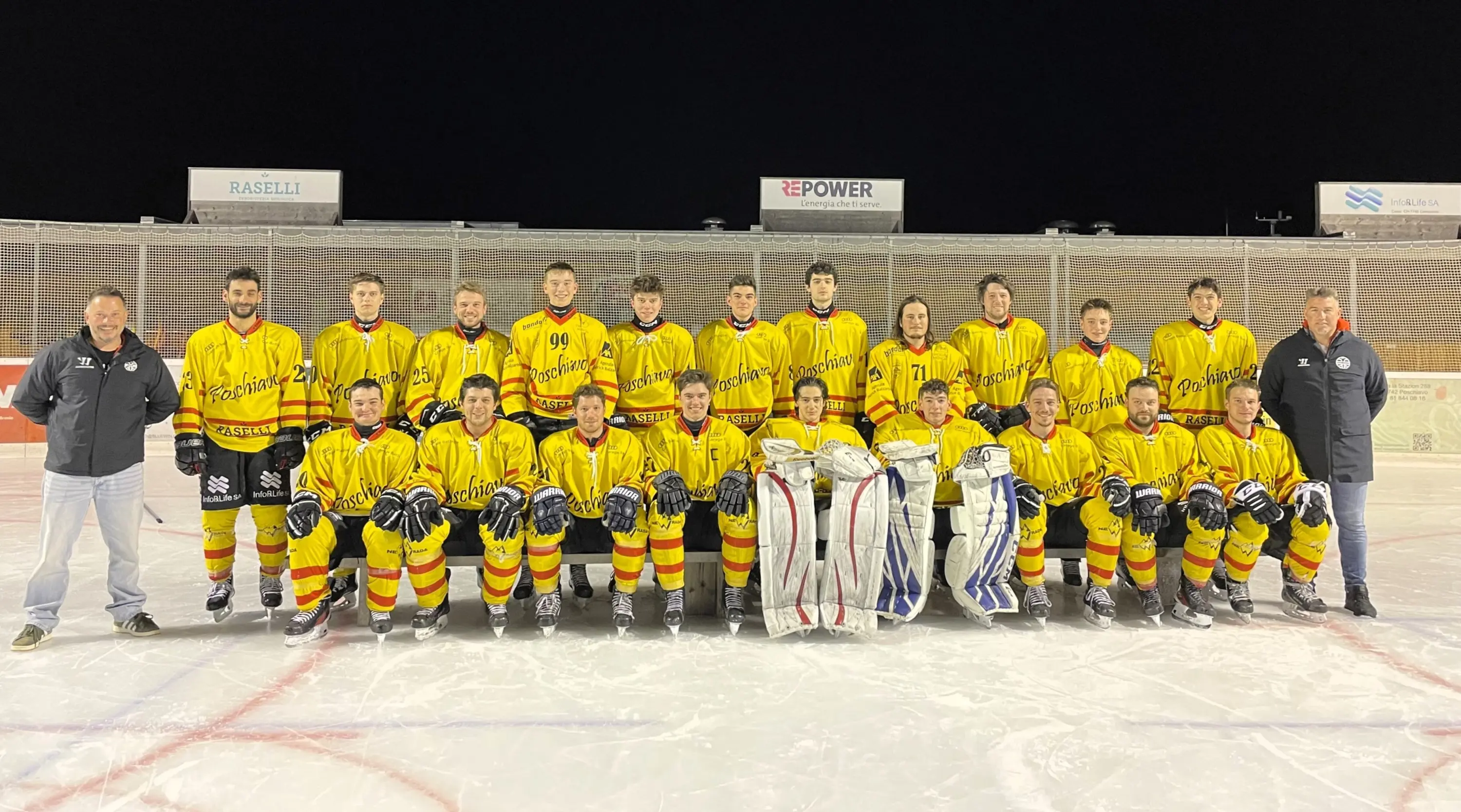 Squadra di hockey su ghiaccio in divisa gialla e rossa, composta da 22 giocatori e due portieri, in posa su un campo di ghiaccio davanti a una struttura di ghiaccio. I membri dello staff sono a sinistra e a destra. È notte e le