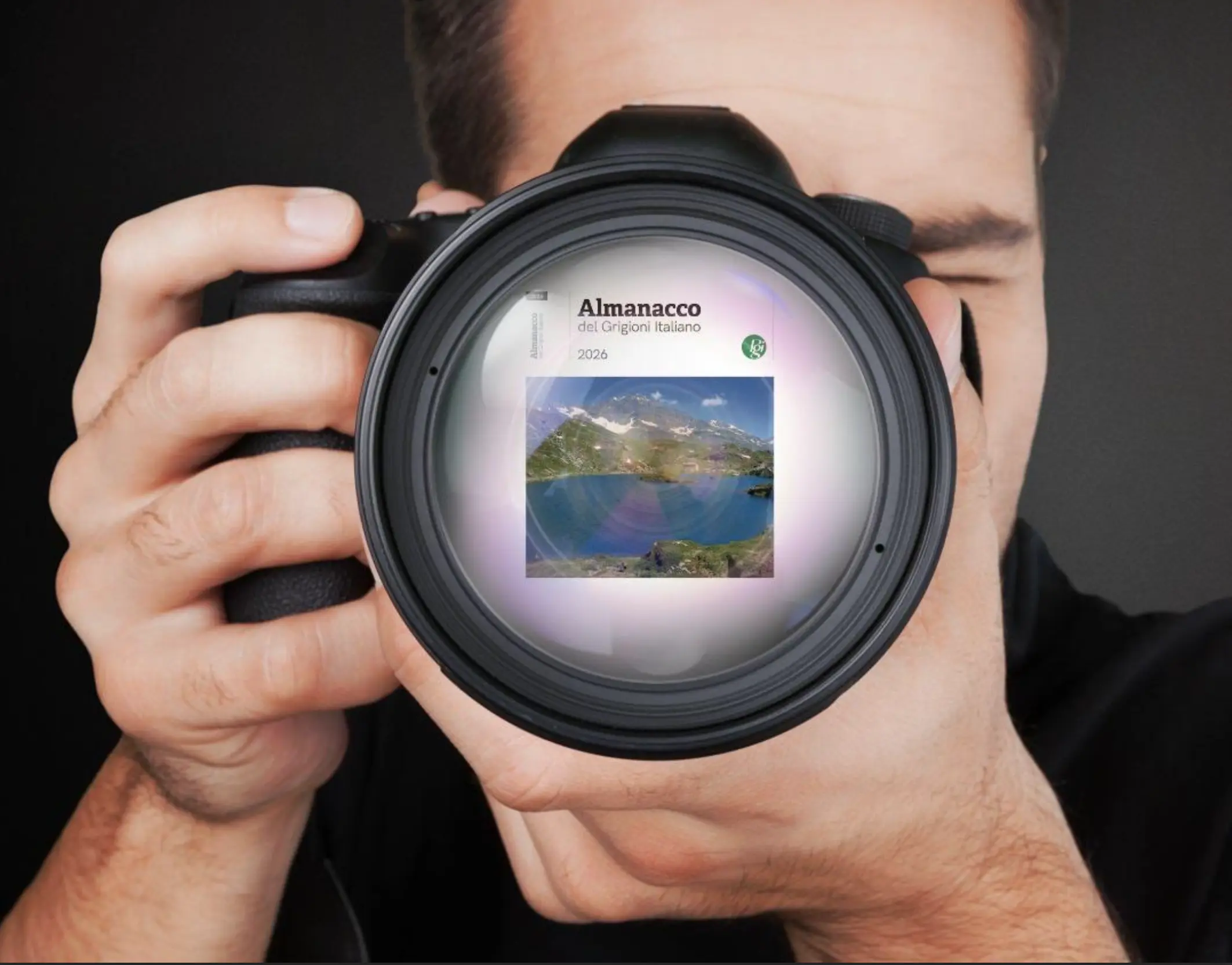 Un fotografo tiene una macchina fotografica con l'obiettivo rivolto verso l'osservatore. Dentro l'obiettivo si vede un'immagine di un paesaggio naturale e una copertina di un calendario chiamato "Almanacco del Grogon italiano 2026".