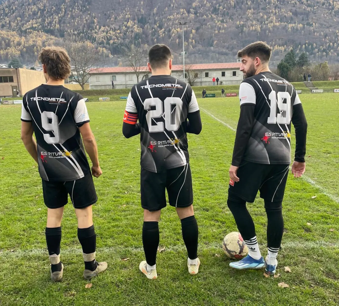 Tre calciatori in divisa nera e bianca, con i numeri 9, 20 e 18 sulla schiena, osservano il campo da gioco. Sullo sfondo si intravedono colline e una struttura sportiva.