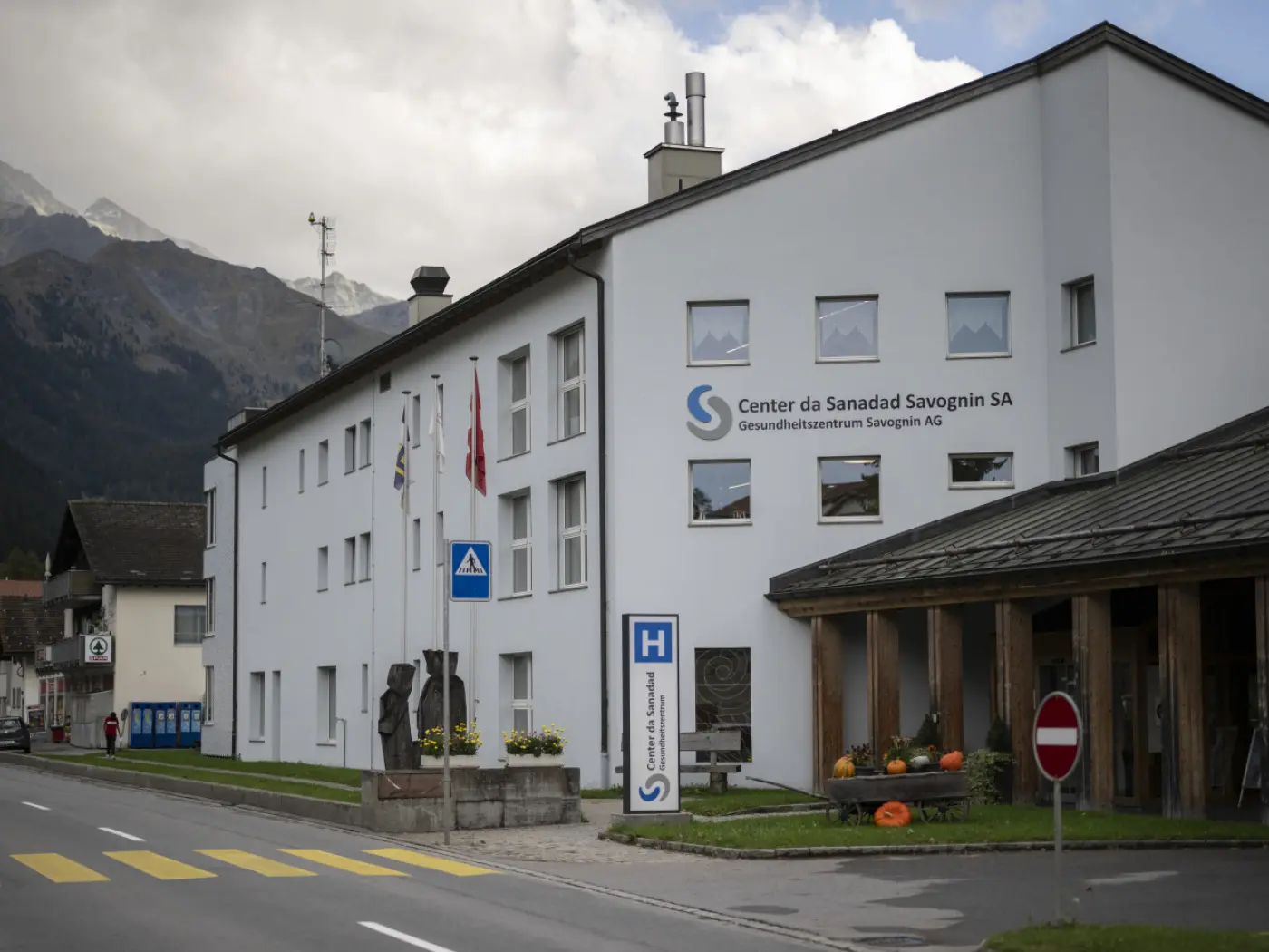 Edificio moderno con il logo "Center da Sanadat Savognin SA". Strada in primo piano con segnali stradali e decorazioni di stagione. Sullo sfondo, montagne e cielo nuvoloso.