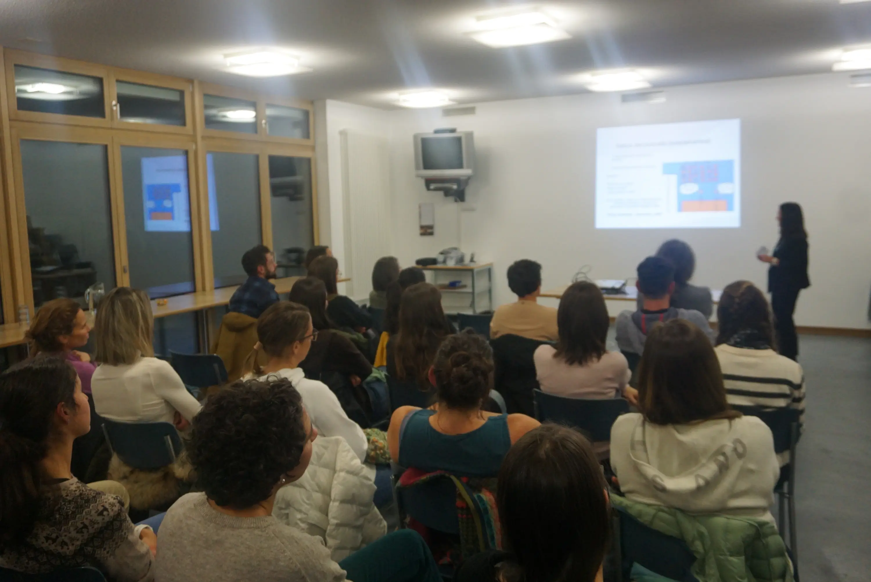 Una sala di formazione con diverse persone sedute che ascoltano un relatore davanti a una presentazione proiettata. La stanza è luminosa e moderna, con un grande schermo e alcune attrezzature tecnologiche visibili.