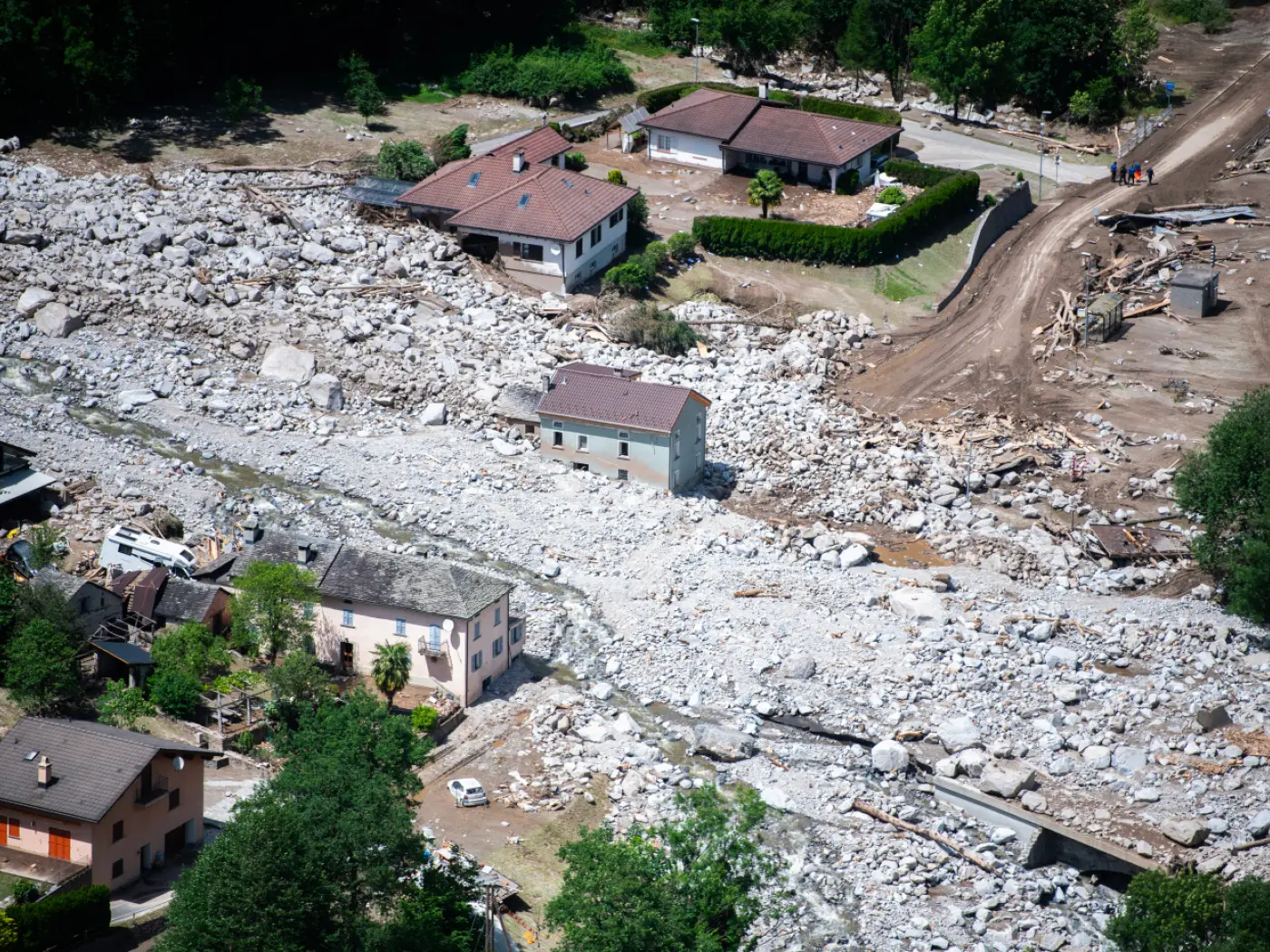 Terreno devastato da un'alluvione, con case circondate da massi e detriti. Alcuni veicoli danneggiati si trovano tra i detriti, mentre un gruppo di persone è visibile in lontananza.