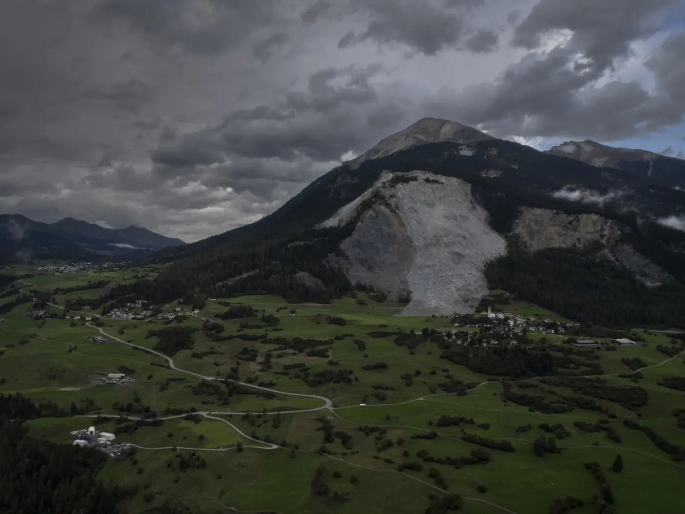 Vista panoramica di una valle montuosa con nuvole scure nel cielo. Un grande masso è scivolato lungo il versante della montagna, mentre il paesaggio