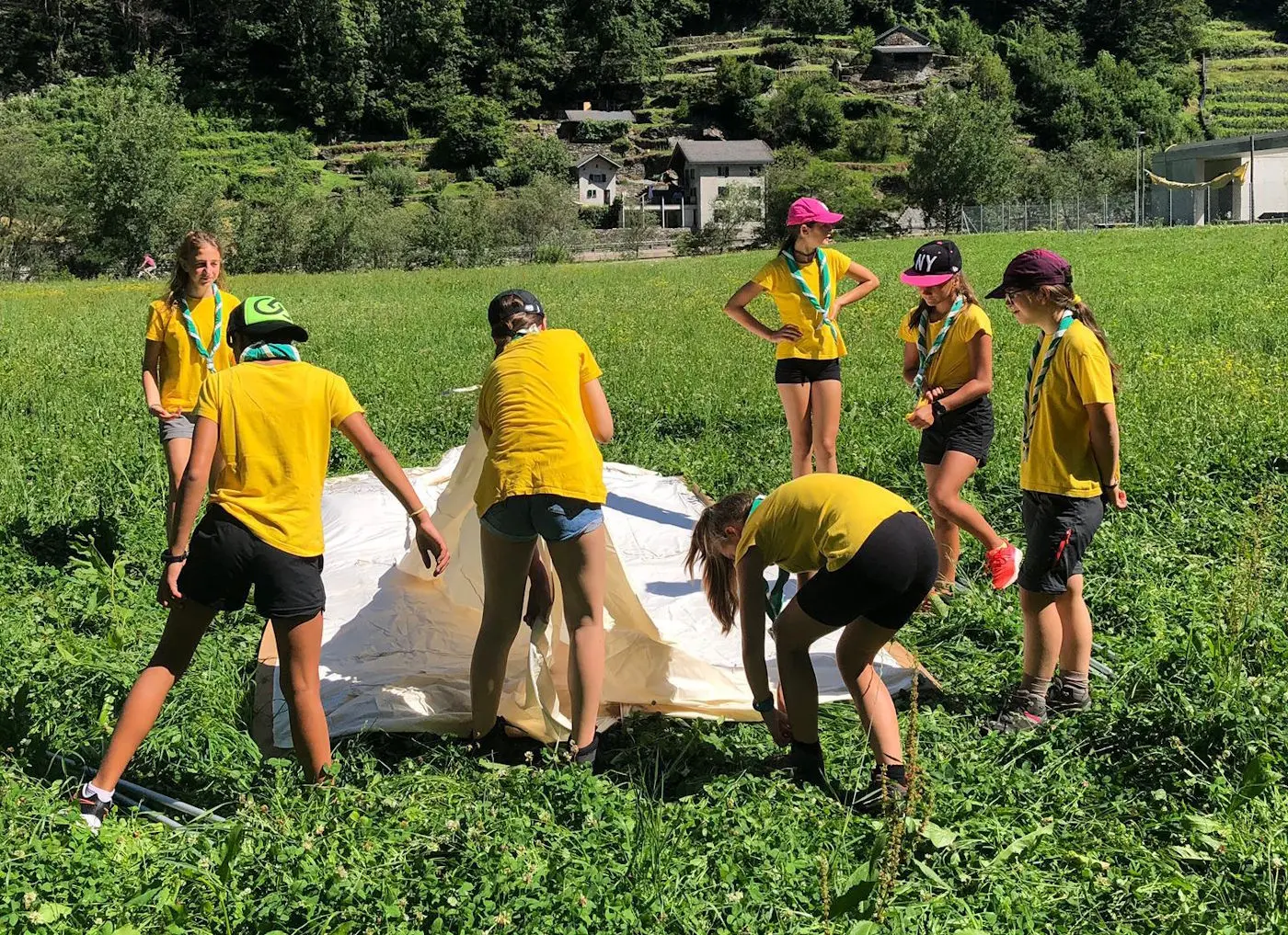 Un gruppo di giovani indossa magliette gialle mentre montano una tenda in un campo verde. Sullo sfondo si vedono case e alberi.
