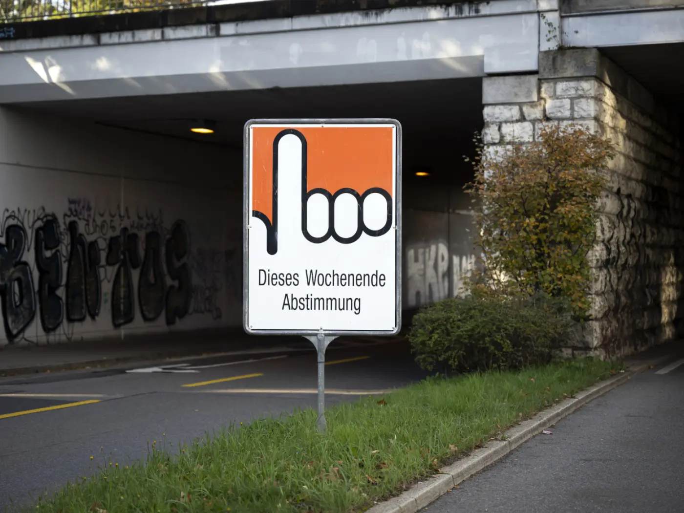 Cartello stradale con un dito indice alzato, accompagnato dal testo "Dieses Wochenende Abstimmung", situato sotto un ponte con graffiti sullo sfondo.