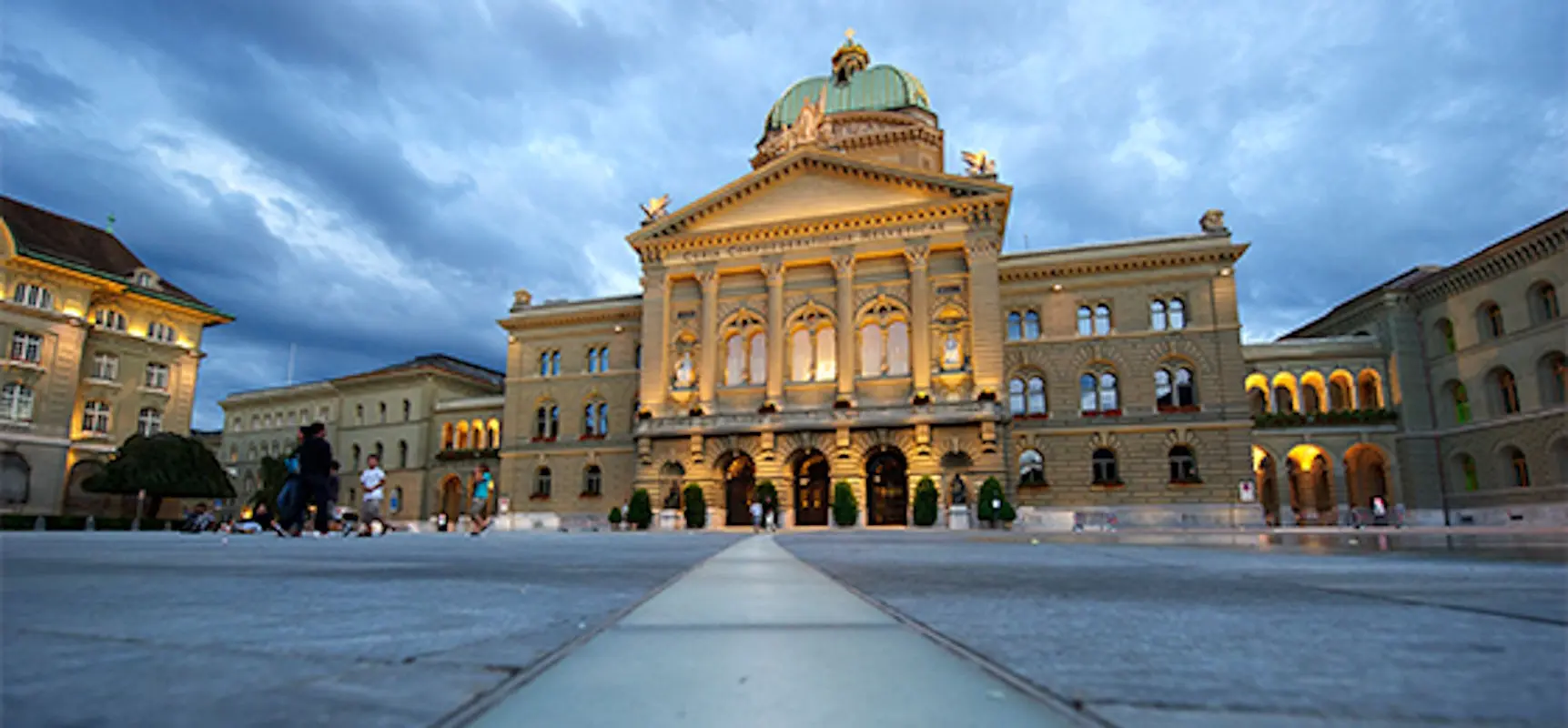 Il Palazzo Federale di Berna, con la sua imponente facciata e la cupola verde, illuminato da luci calde al tramonto. Nuvole grigie si stag