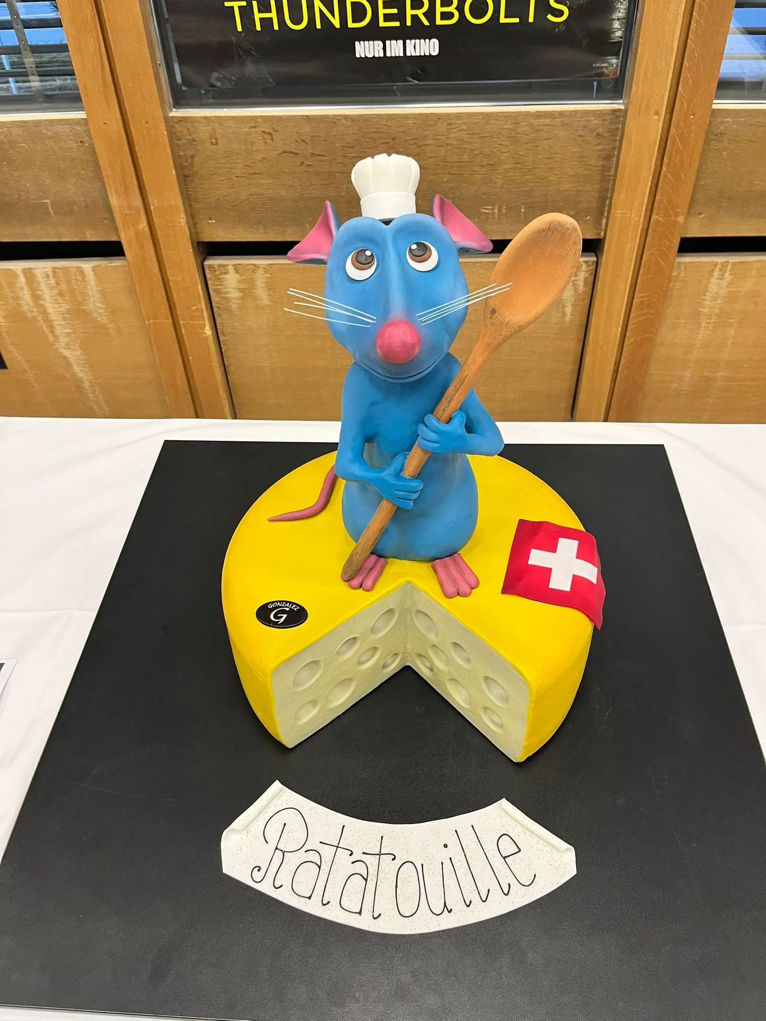 Una torta decorata che rappresenta un topo con un cappello da cuoco, che tiene un cucchiaio, sopra un grande formaggio giallo con buchi e una bandiera