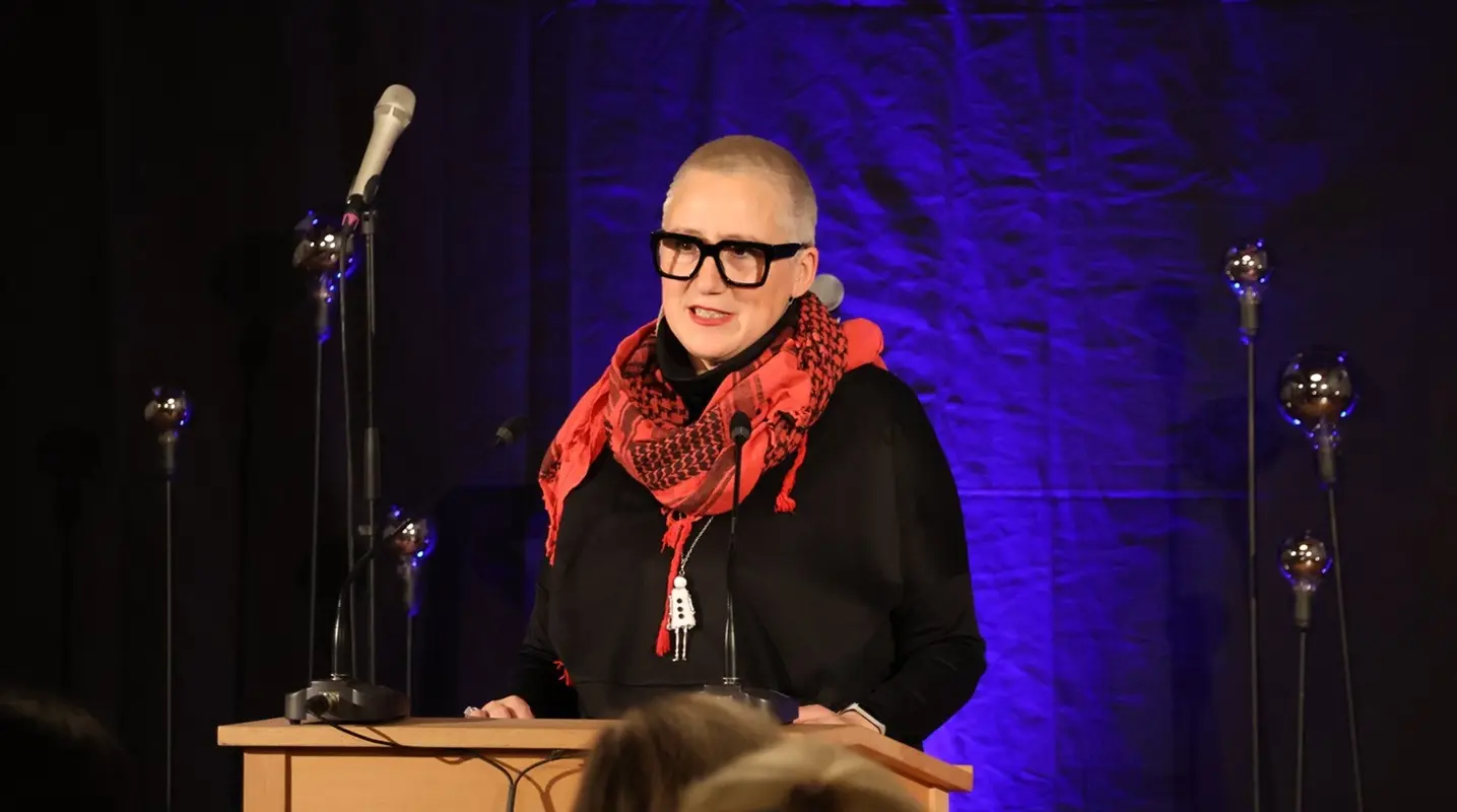 Una donna con occhiali grandi e capelli rasati parla a un podio, indossando una sciarpa rossa su uno sfondo blu e nero. Un microfono è posiz