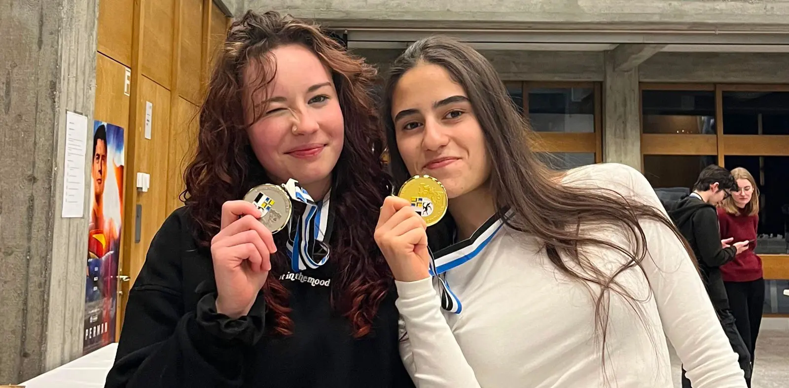 Due ragazze sorridenti mostrano le loro medaglie. Una tiene una medaglia d'argento, l'altra una medaglia d'oro. Sullo sfondo si intravedono