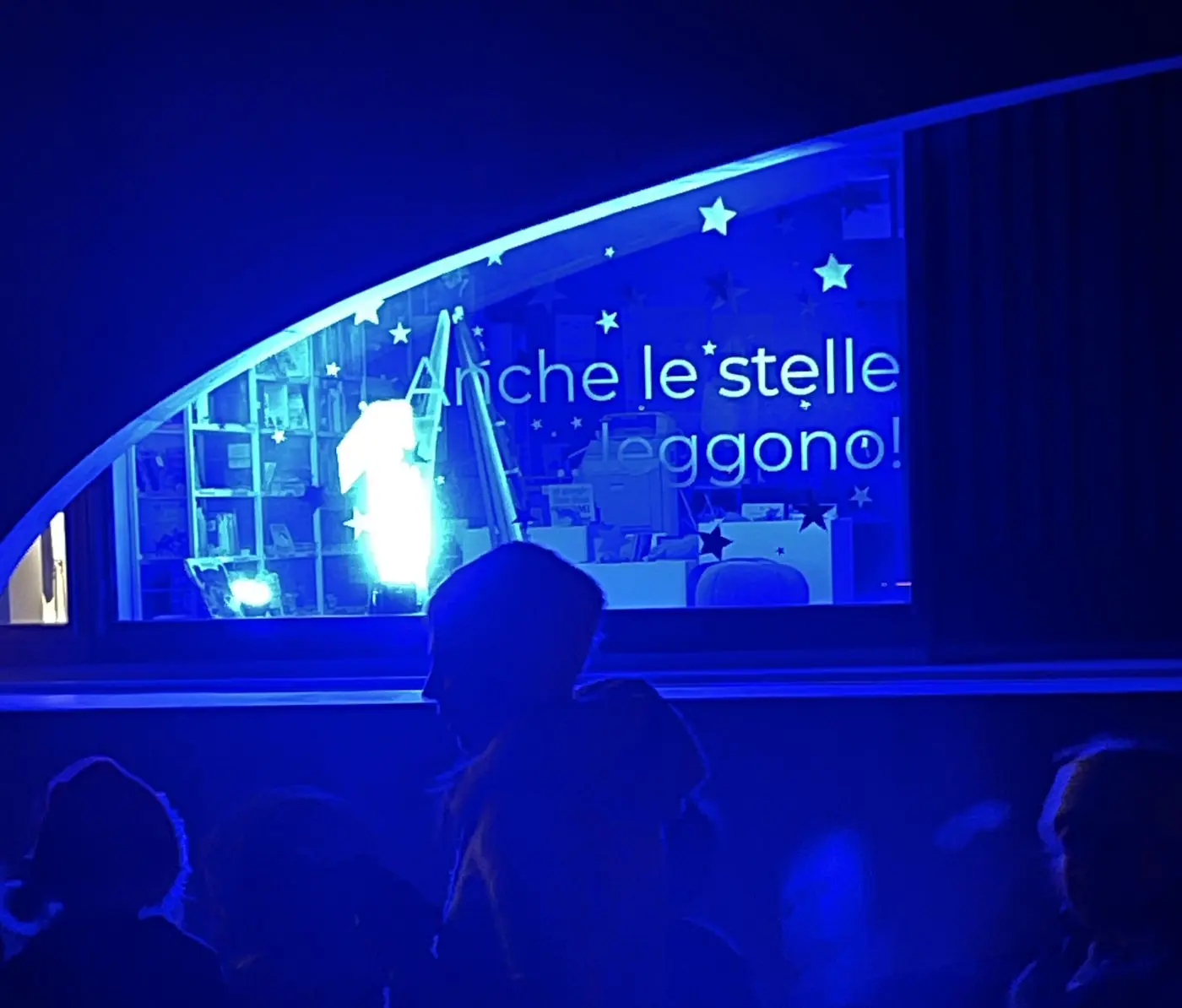 Finestra illuminata con scritto "Anche le stelle leggono!" e decorazioni stellari, silhouette di persone in colloquio all'esterno.