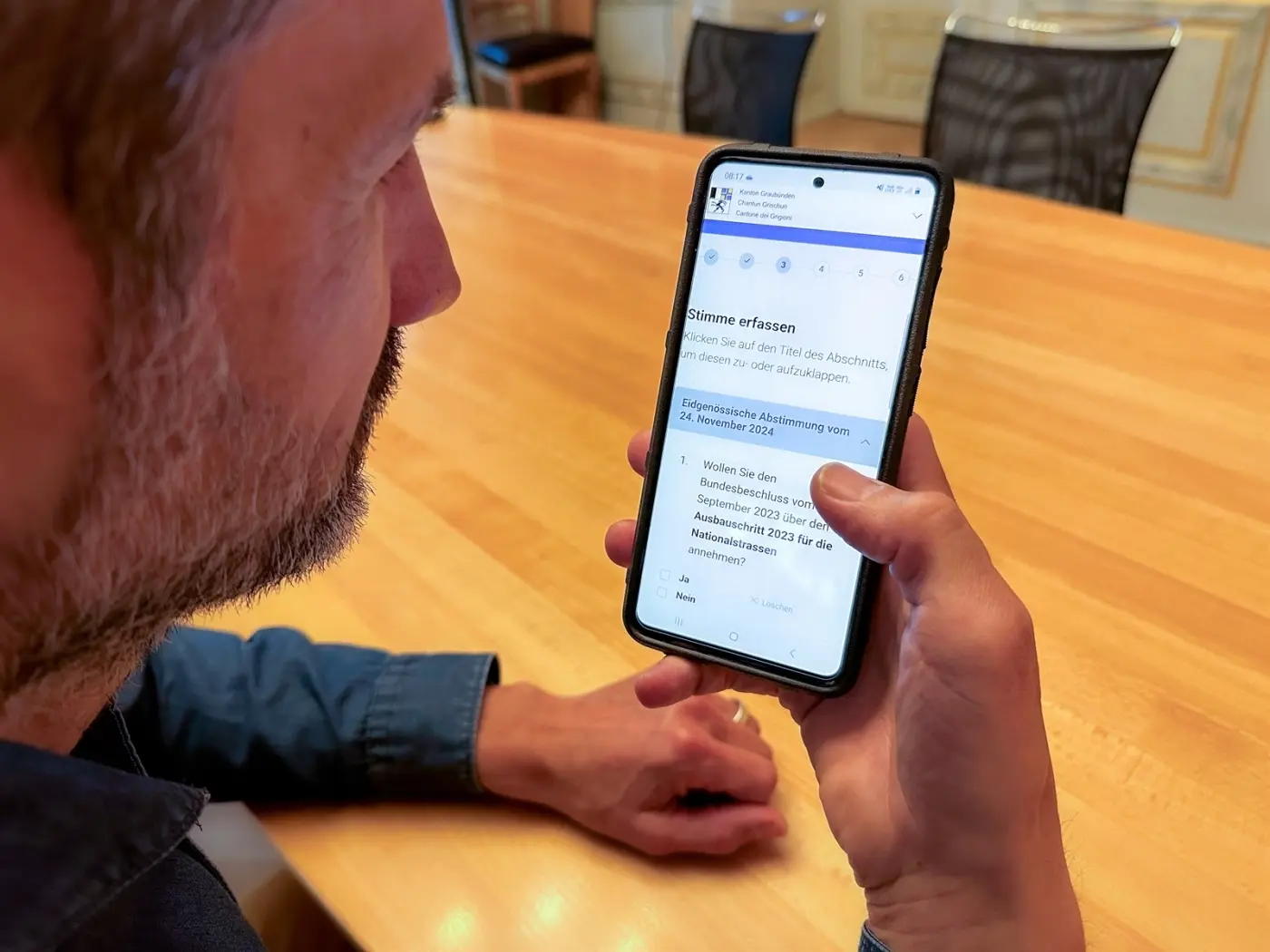 Un uomo tiene in mano uno smartphone, leggendo un documento sullo schermo. È seduto a un tavolo di legno in un ambiente d'ufficio.