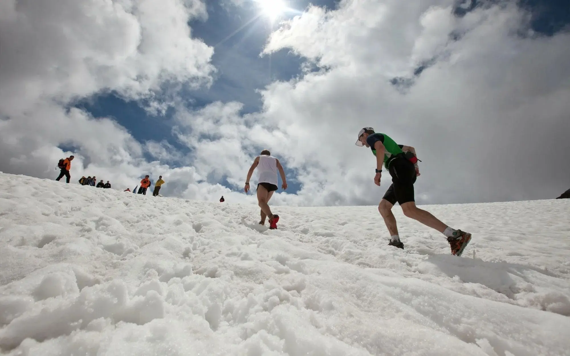 Due corridori salgono su un pendio innevato, sotto un cielo parzialmente nuvoloso. Sullo sfondo, altre persone osservano o partecipano all'attività.