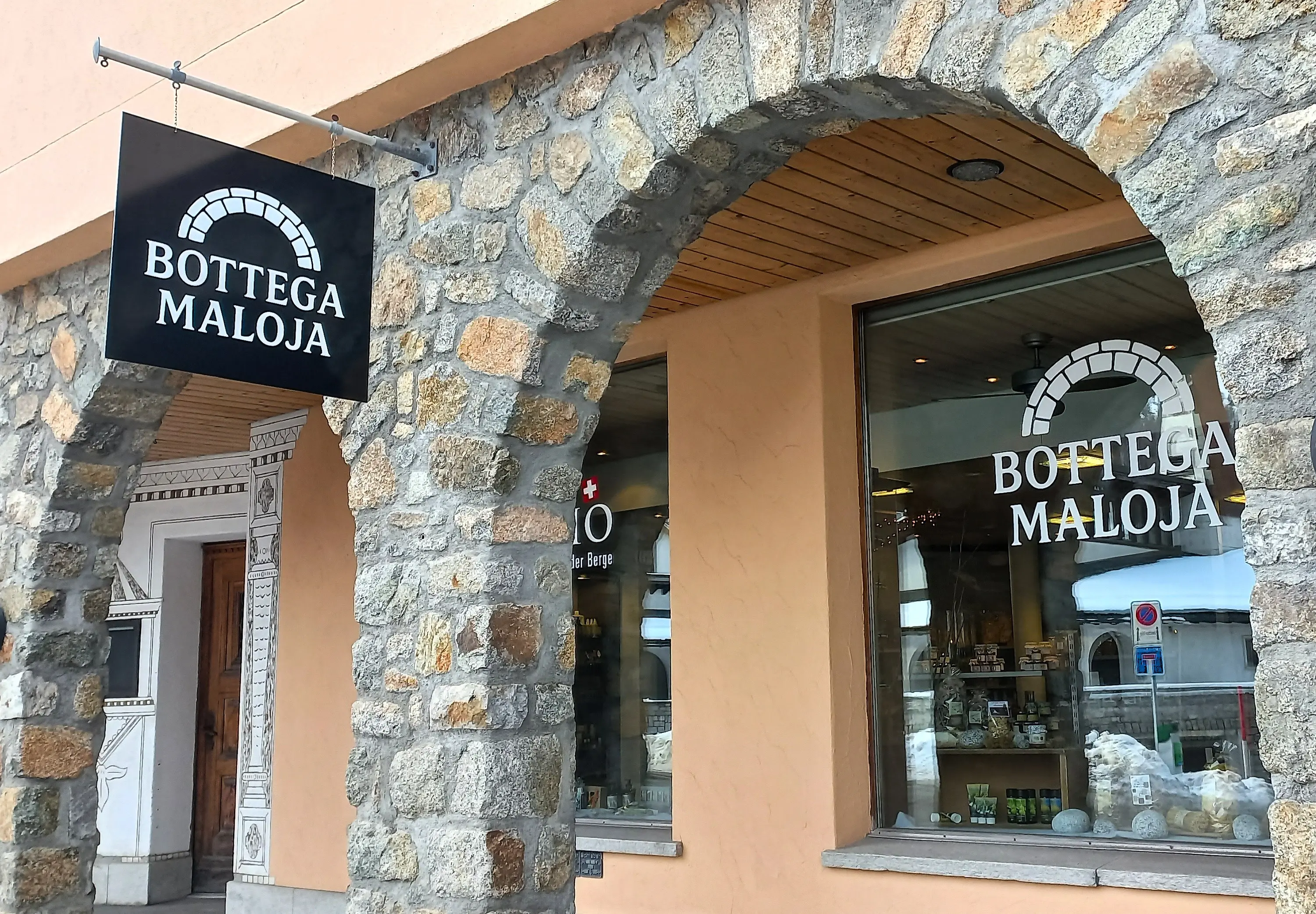 Insegna di legno con la scritta "Bottega Maloja" sopra l'entrata di un negozio con finestre che mostrano prodotti esposti. La struttura è caratter