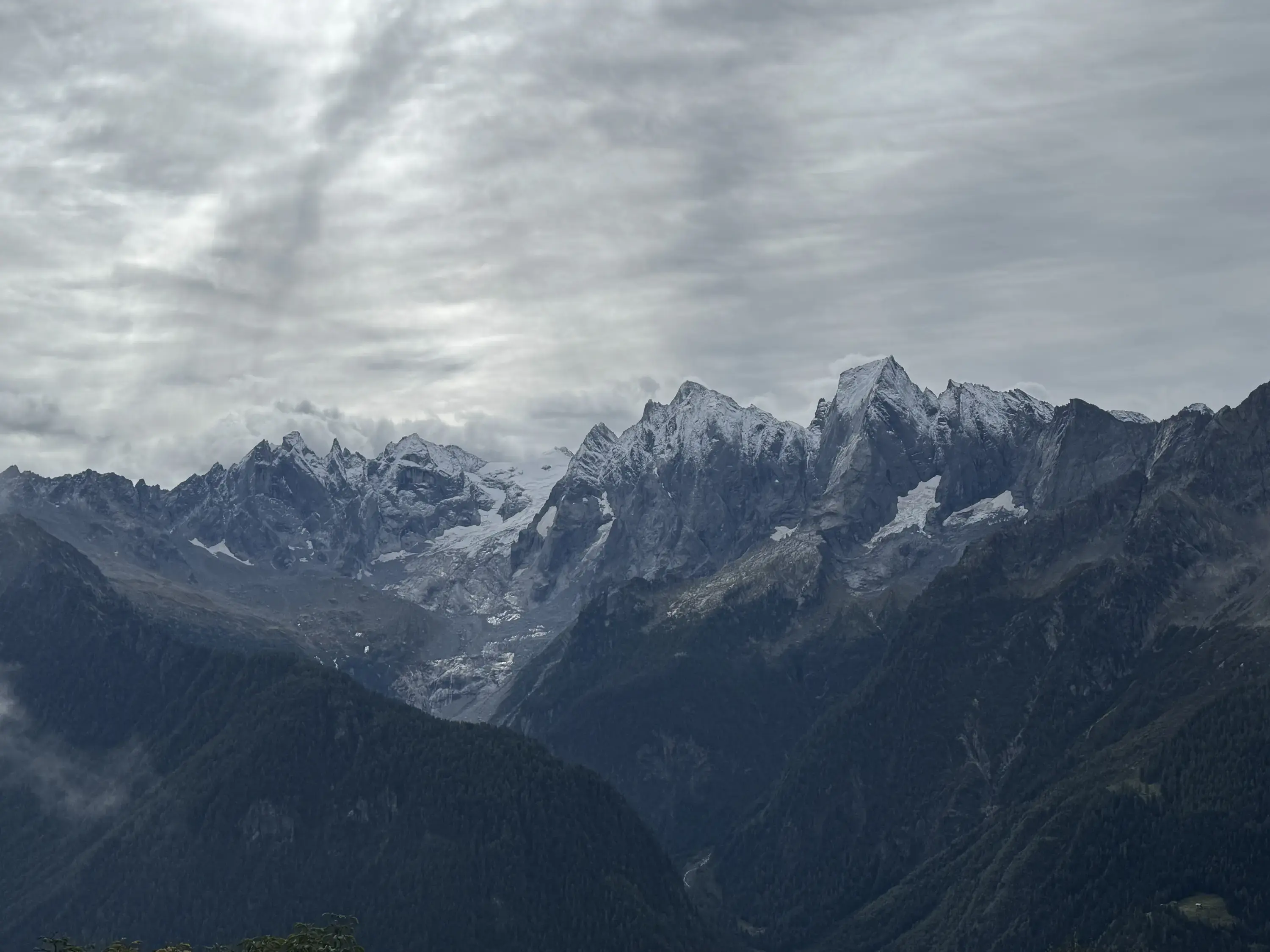 Maestose montagne coperte di neve sullo sfondo di un cielo nuvoloso. La vallata sottostante &egrave; ricoperta di vegetazione verde scuro.