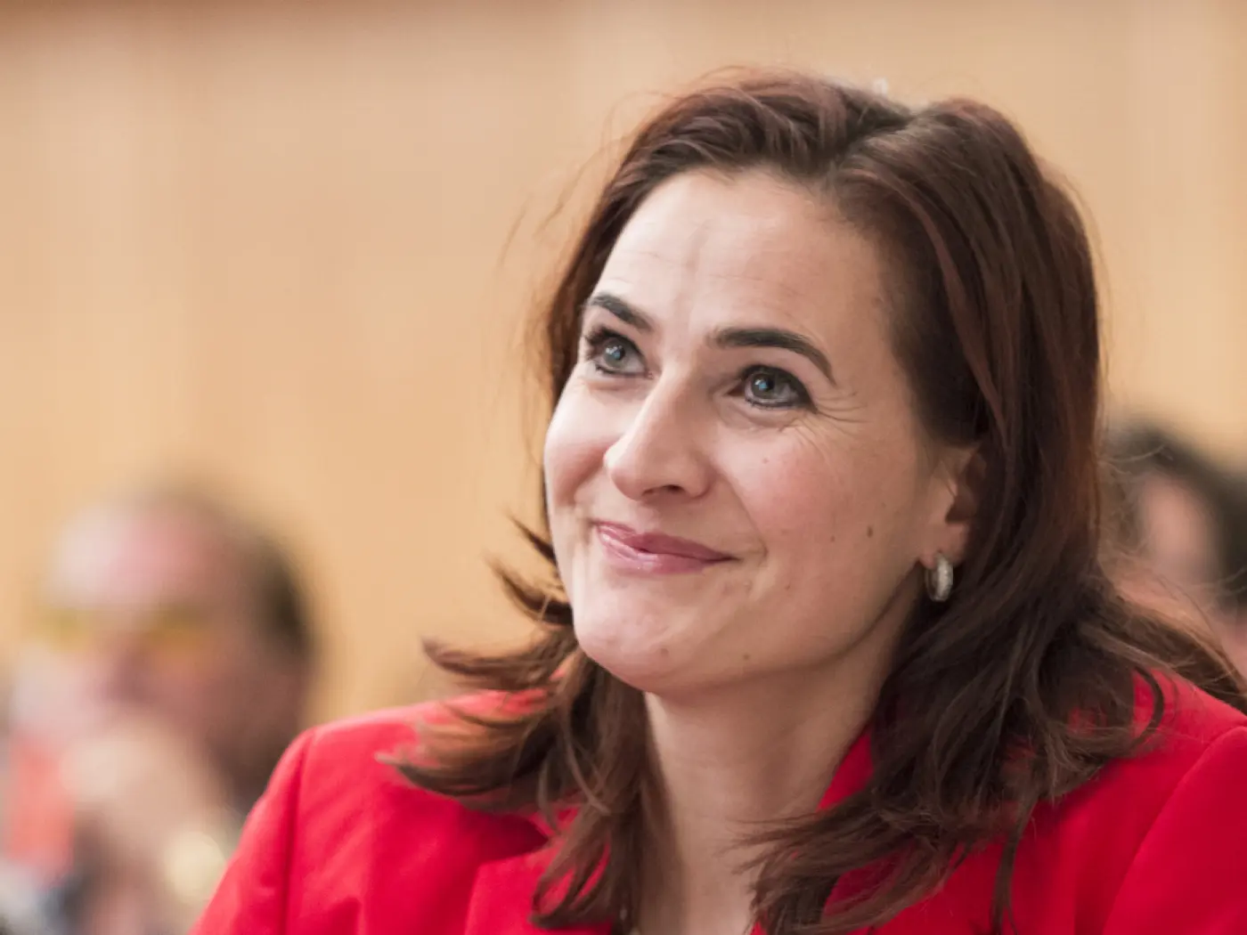 Donna sorridente con capelli castani e lunghi, indossa una giacca rossa. Lo sfondo mostra un ambiente di riunione con altri partecipanti in lontananza.