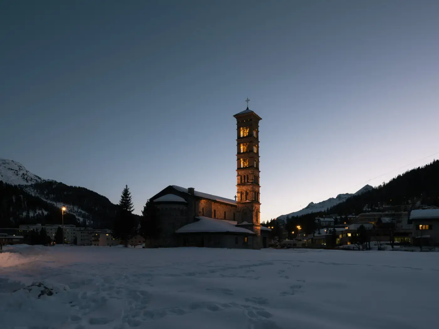 Un'antica chiesa con un campanile alto illuminato, circondata da un paesaggio innevato al crepuscolo. Sullo sfondo si vedono le montagne.