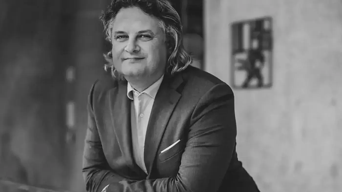 Un uomo con capelli lunghi e ricci indossa un completo elegante mentre posa con le braccia incrociate, sorridendo leggermente. Sullo sfondo si intravede un