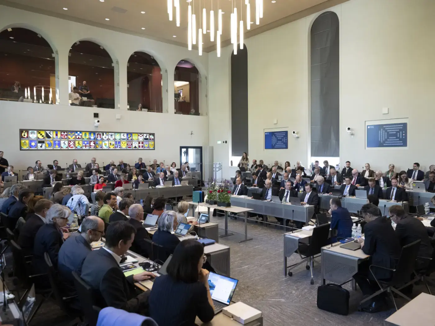 Una sala conferenze affollata con partecipanti seduti a tavoli disposti in modo ordinato. Al centro, il pubblico ascolta relatori su un palco. Luci moderne e opere
