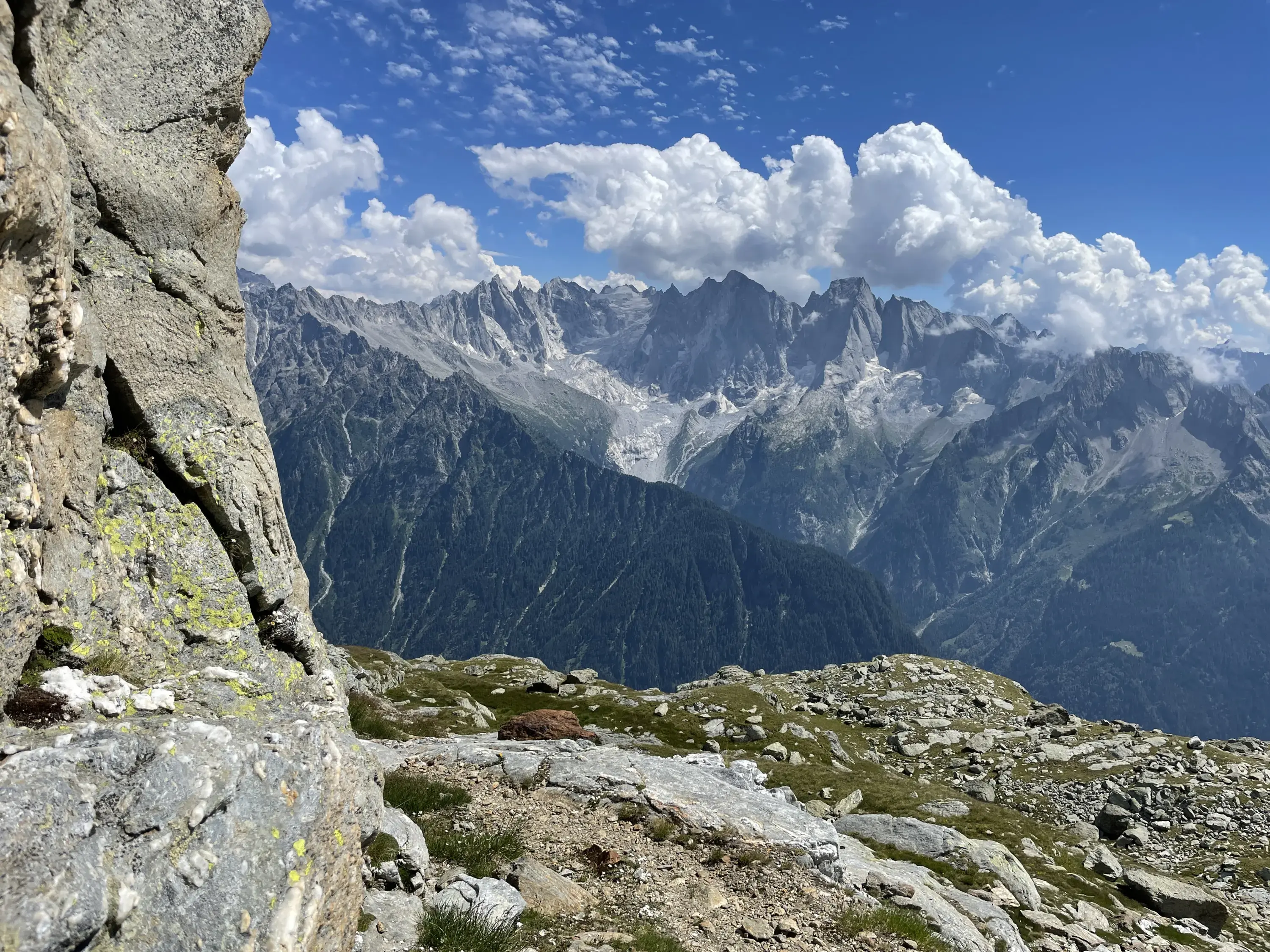 Panorama montano con vette imponenti, nuvole bianche e cielo blu, visto da una roccia in primo piano.