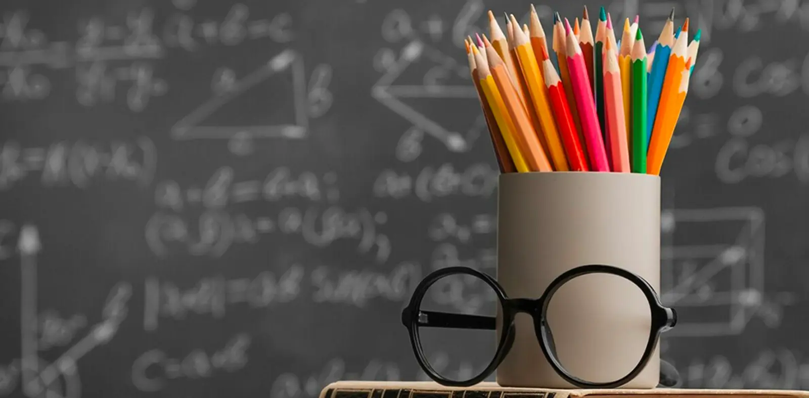 Bicchierino di matite colorate su un tavolo con occhiali neri in primo piano, sullo sfondo una lavagna con formule matematiche.