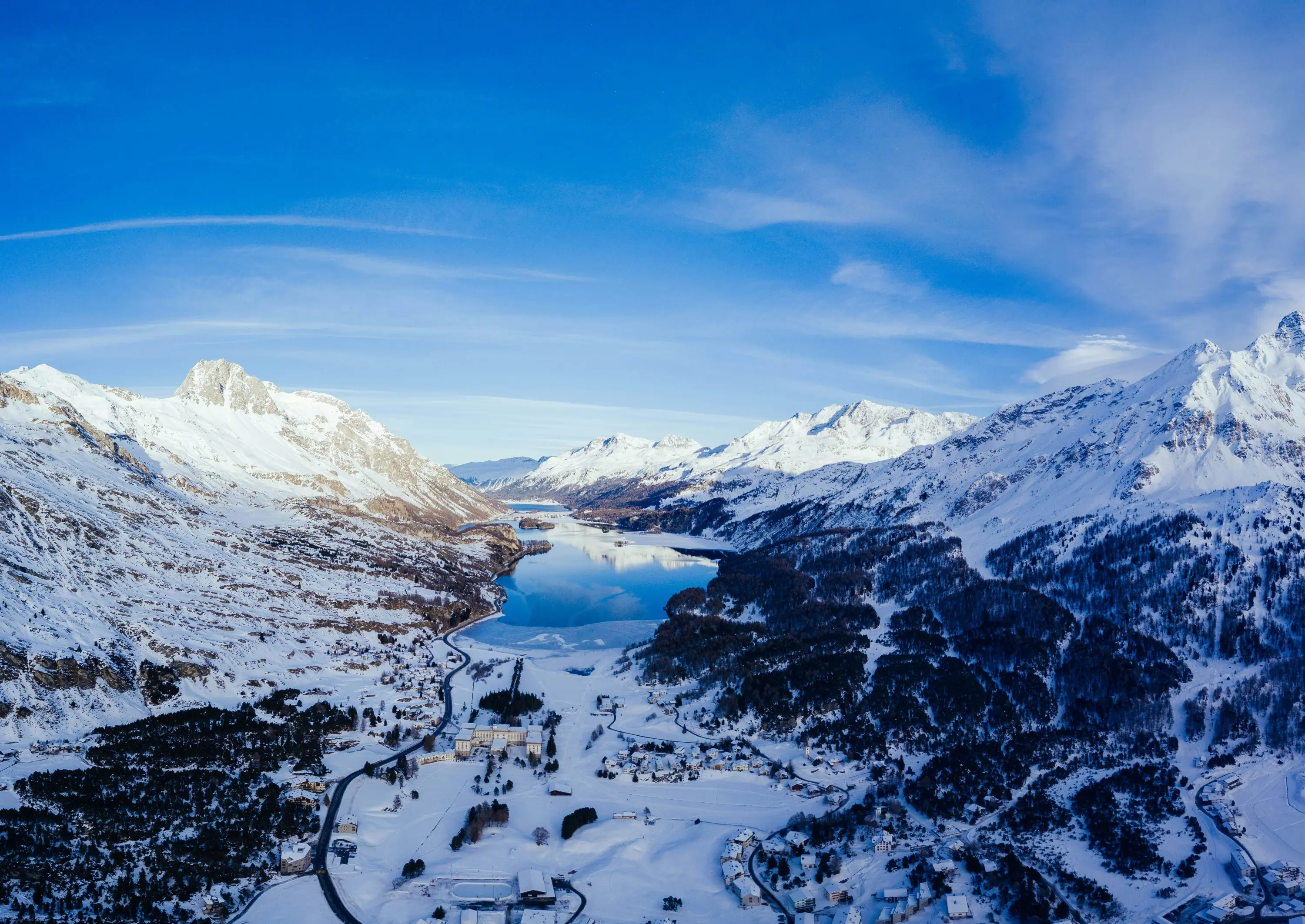 Un paesaggio montano innevato con un lago sereno circondato da vette maestose. Il cielo blu chiaro è attraversato da nuvole leggere, mentre i villaggi e le foreste si estendono lungo le rive del lago.