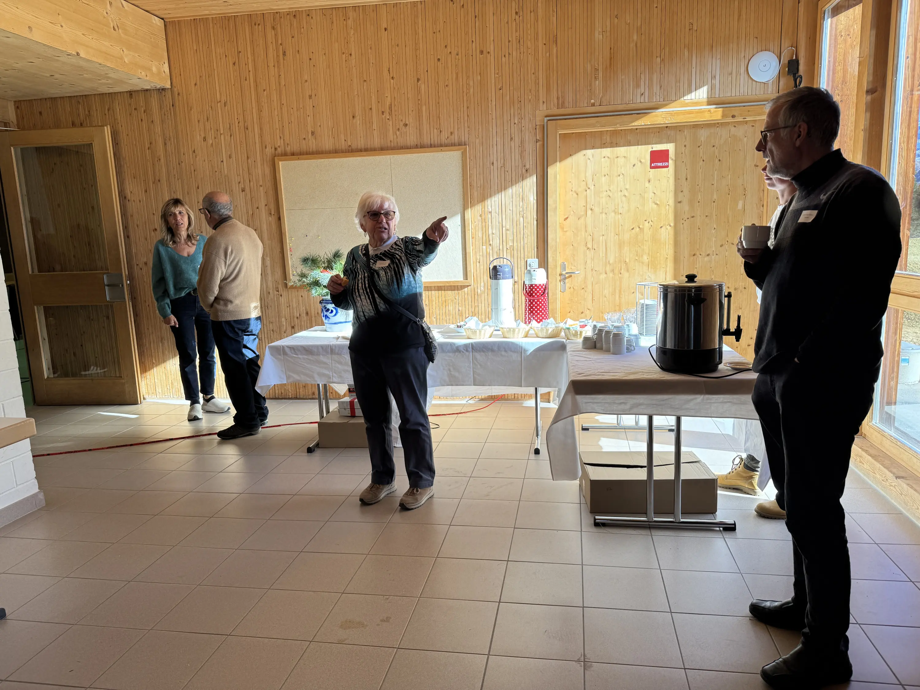 Un gruppo di persone in una stanza con pareti in legno, una donna anziana indica qualcosa mentre parla. Al tavolo è disposto un buffet con bevande e snack. Altre persone, alcune in piedi e altre sedute, osservano la scena.