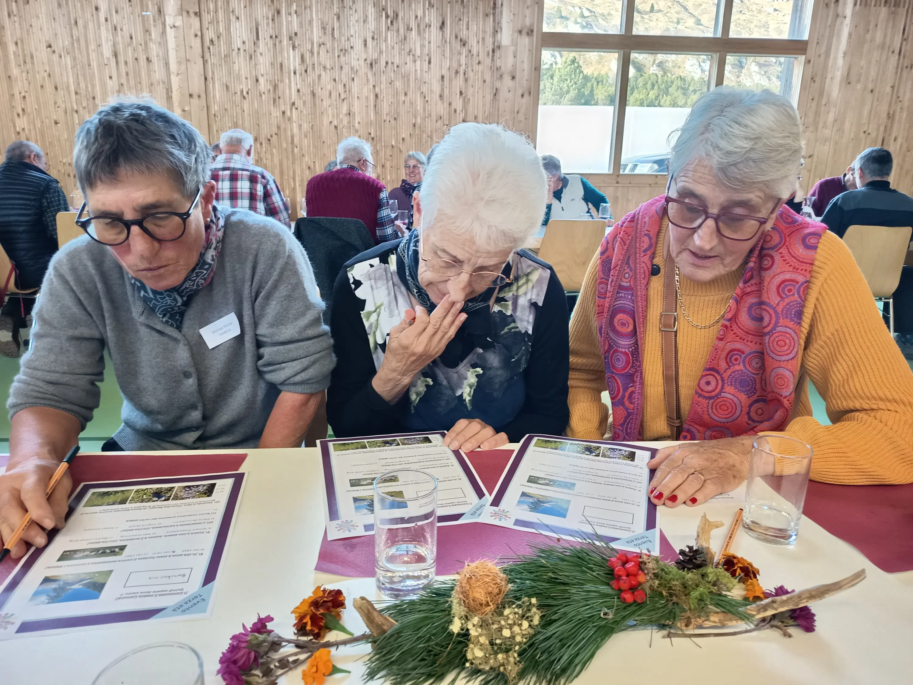 Tre donne anziane siedono attorno a un tavolo, esaminando documenti con immagini. Due di loro indossano occhiali e mostrano espressioni concentrate, mentre una terza sembra pensierosa. Sul tavolo ci sono bicchieri d'acqua e decor