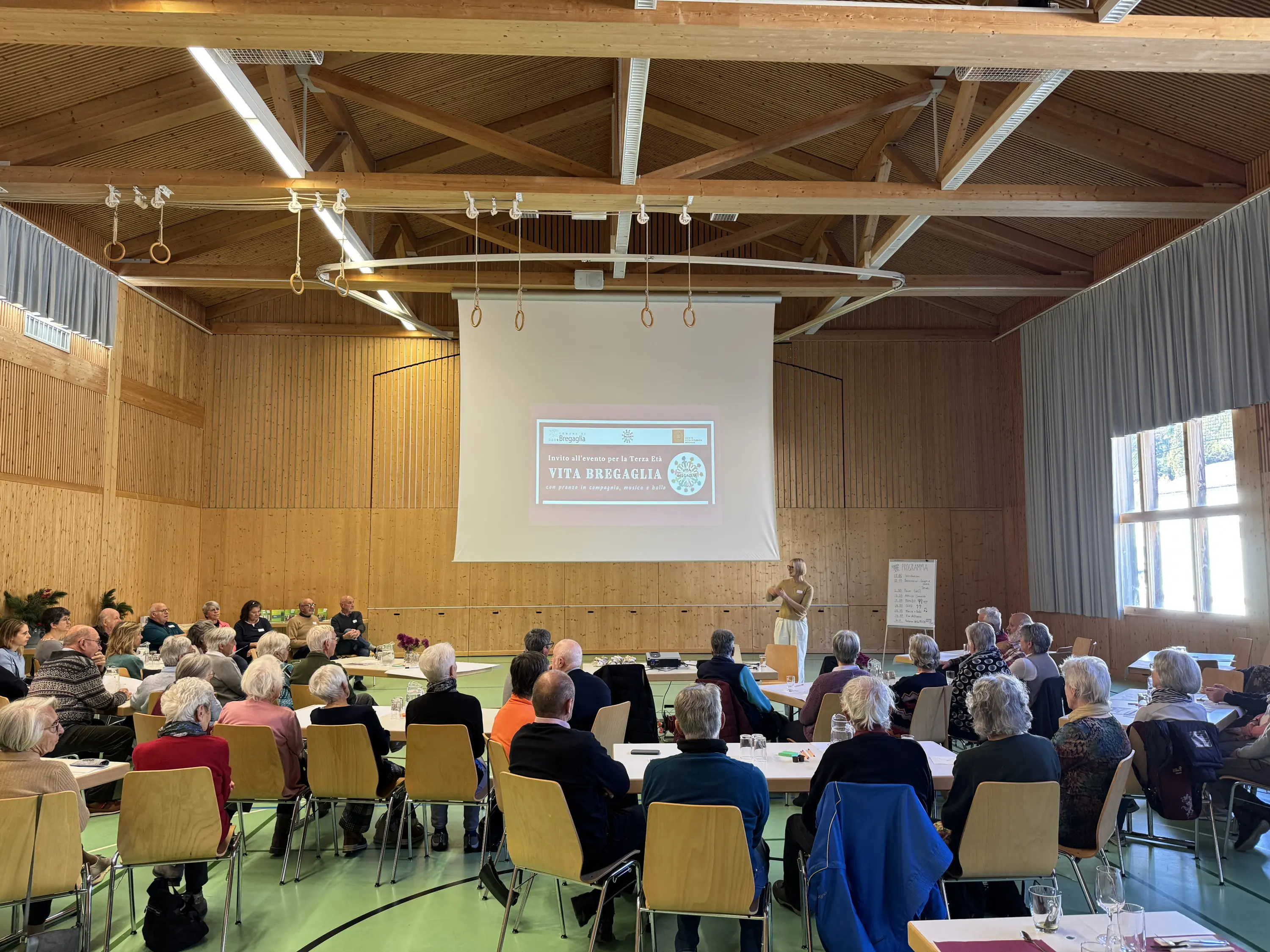Un gruppo di persone anziane partecipa a una conferenza in una sala con soffitto in legno. Sullo schermo è visibile un titolo che recita "Vita e Brigaglie". I partecipanti sono seduti a tavoli, concentrati sul relatore.