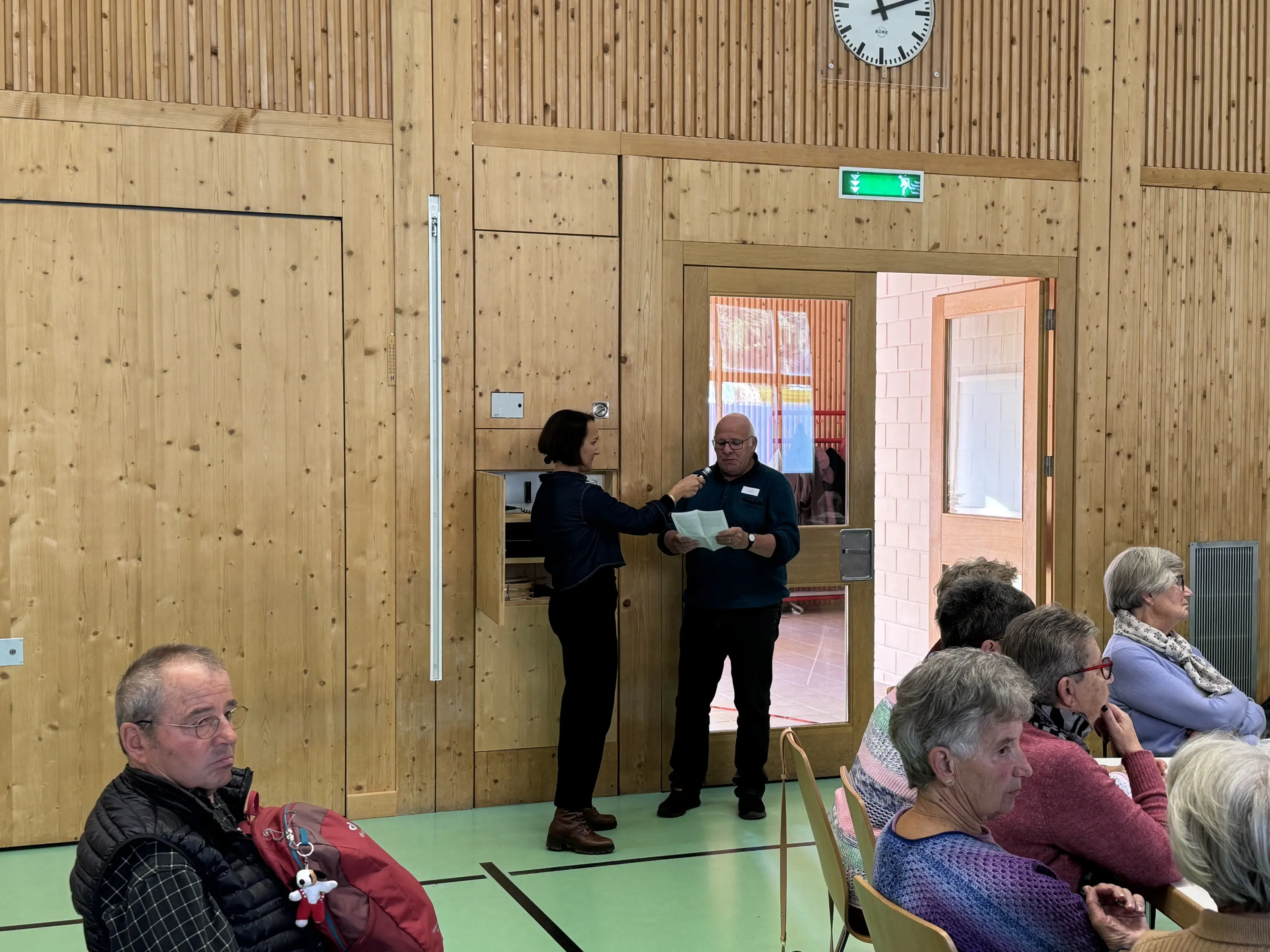 Due persone in un ambiente interno di legno. Una donna porge un documento a un uomo in un pannello di vetro. Dietro di loro, un gruppo di persone sedute osserva. Una grande finestra illumina la stanza.