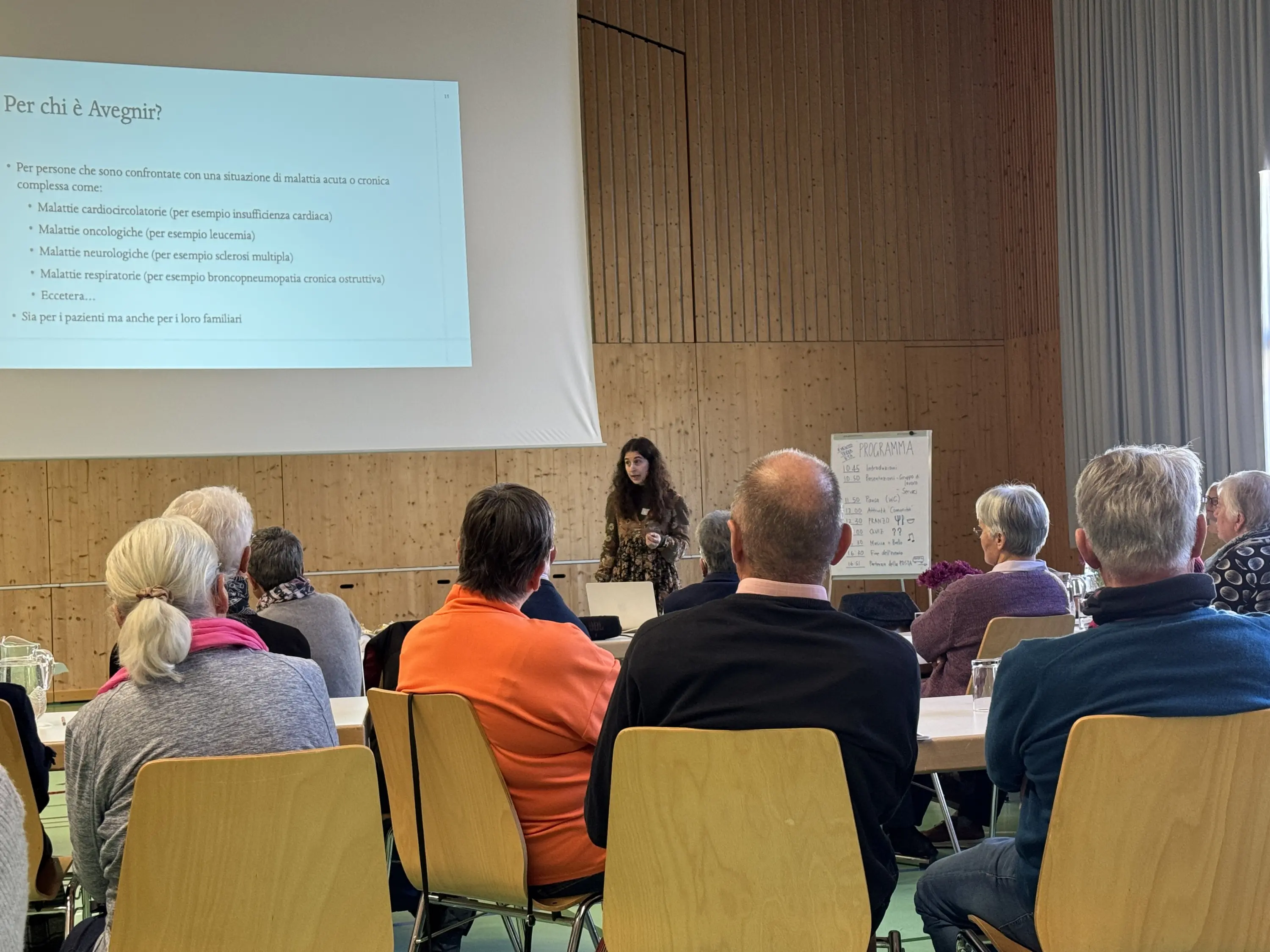 Una presentazione in corso con una relatrice che parla a un pubblico di adulti seduti. Sullo schermo dietro di lei si vedono delle informazioni scritte riguardo a un tema. L'ambiente è luminoso e scarno, con sedie e un tavolo.