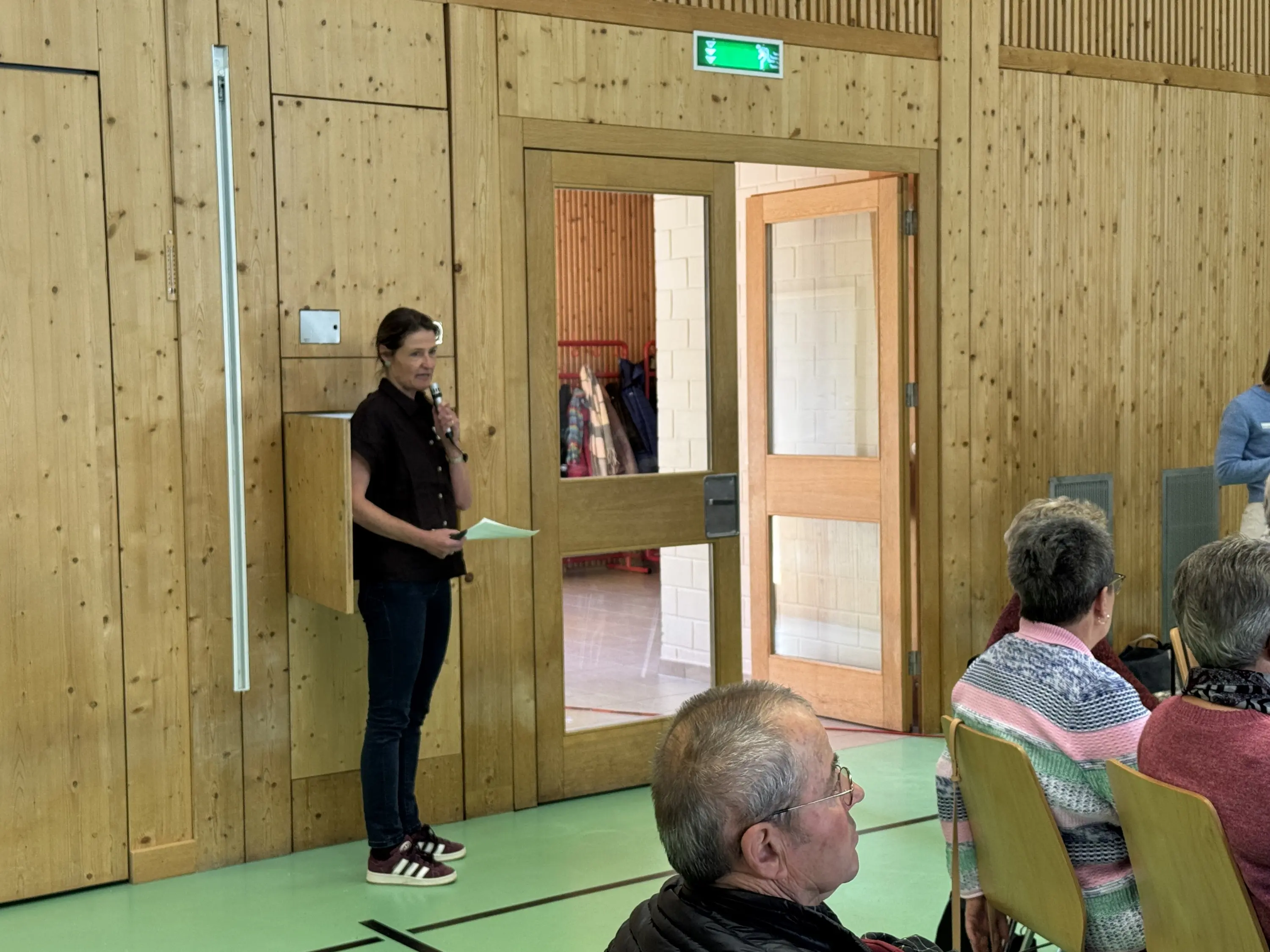 Una persona in piedi accanto a una porta, che tiene un foglio in mano, mentre un gruppo di persone è seduto in una sala con pareti in legno.