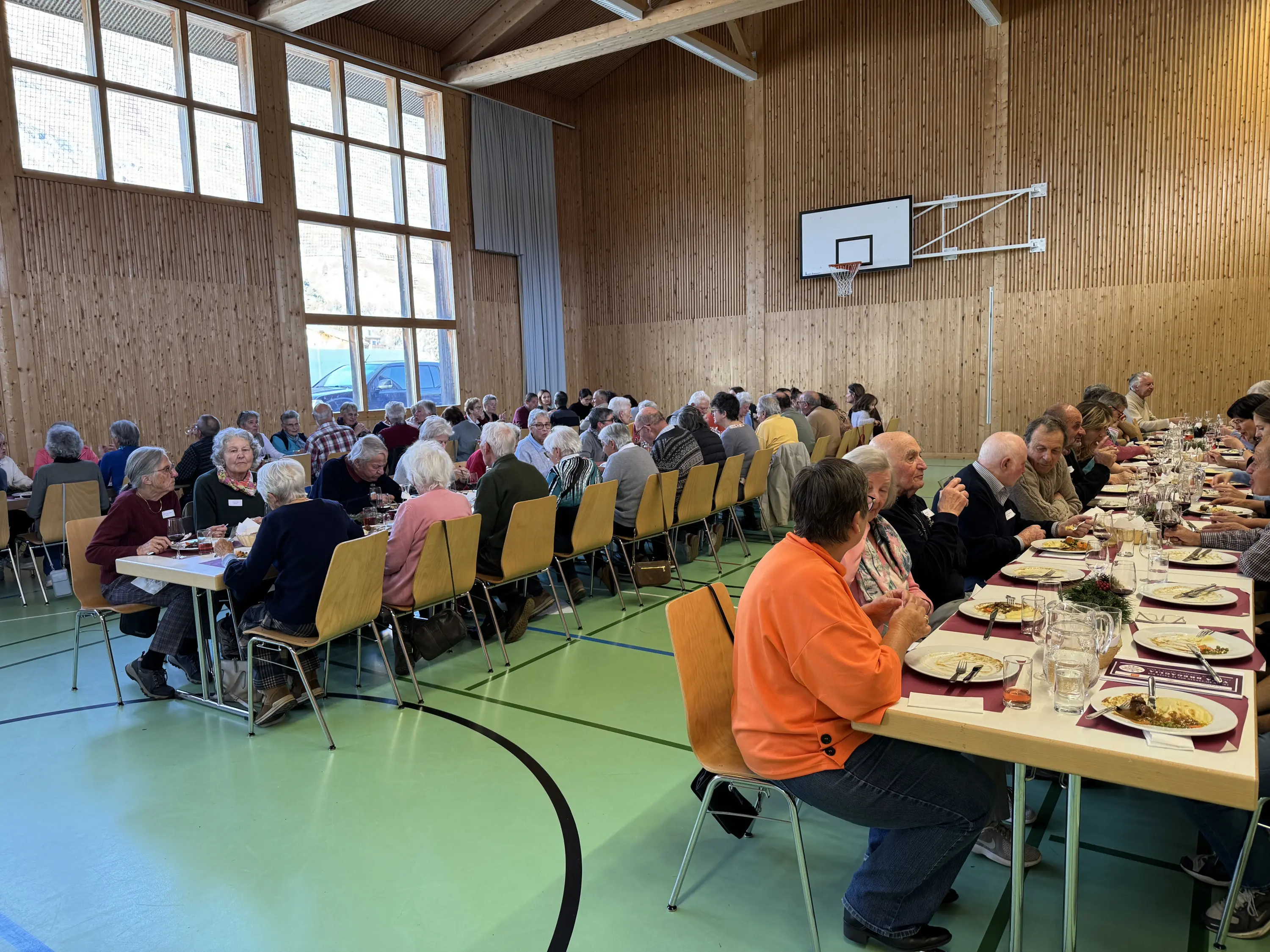 Un gruppo di persone anziane si riunisce in una sala luminosa per un pranzo comunitario. I partecipanti sono seduti a lunghe tavolate, condividendo cibo e conversazione in un ambiente accogliente con pareti di legno.