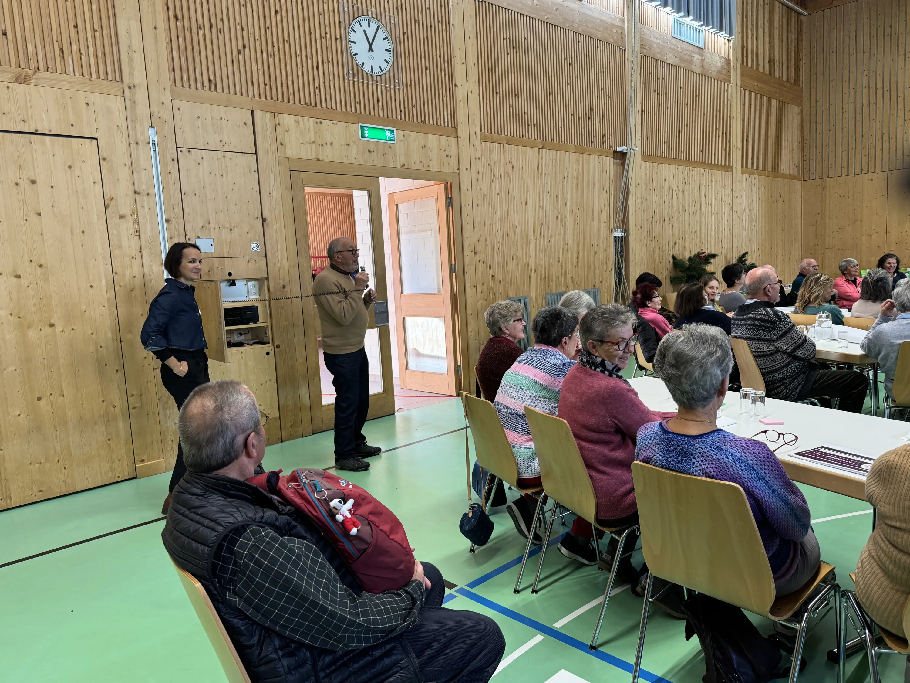 Un gruppo di persone sedute in una sala con pareti di legno. Un uomo anziano sta parlando in piedi davanti a un pubblico attento, mentre un'altra persona, anch'essa in piedi, osserva. Tutti i partecipanti sembrano coinvolti nella discussione. Sul