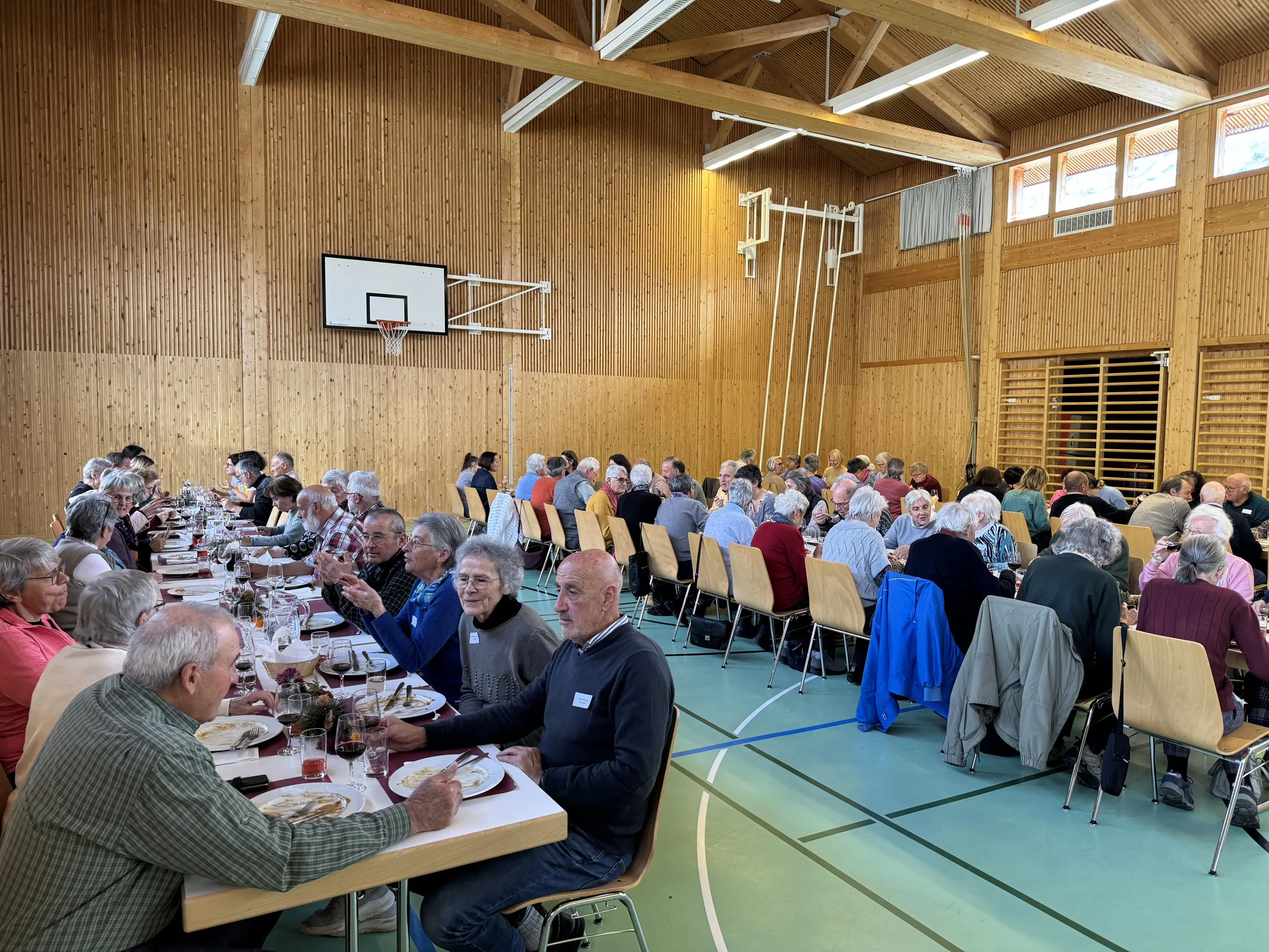 Una sala con molte persone anziane sedute a lungo a un tavolo, pronte per un pranzo. Le pareti in legno e l'illuminazione naturale creano un'atmosfera accogliente.
