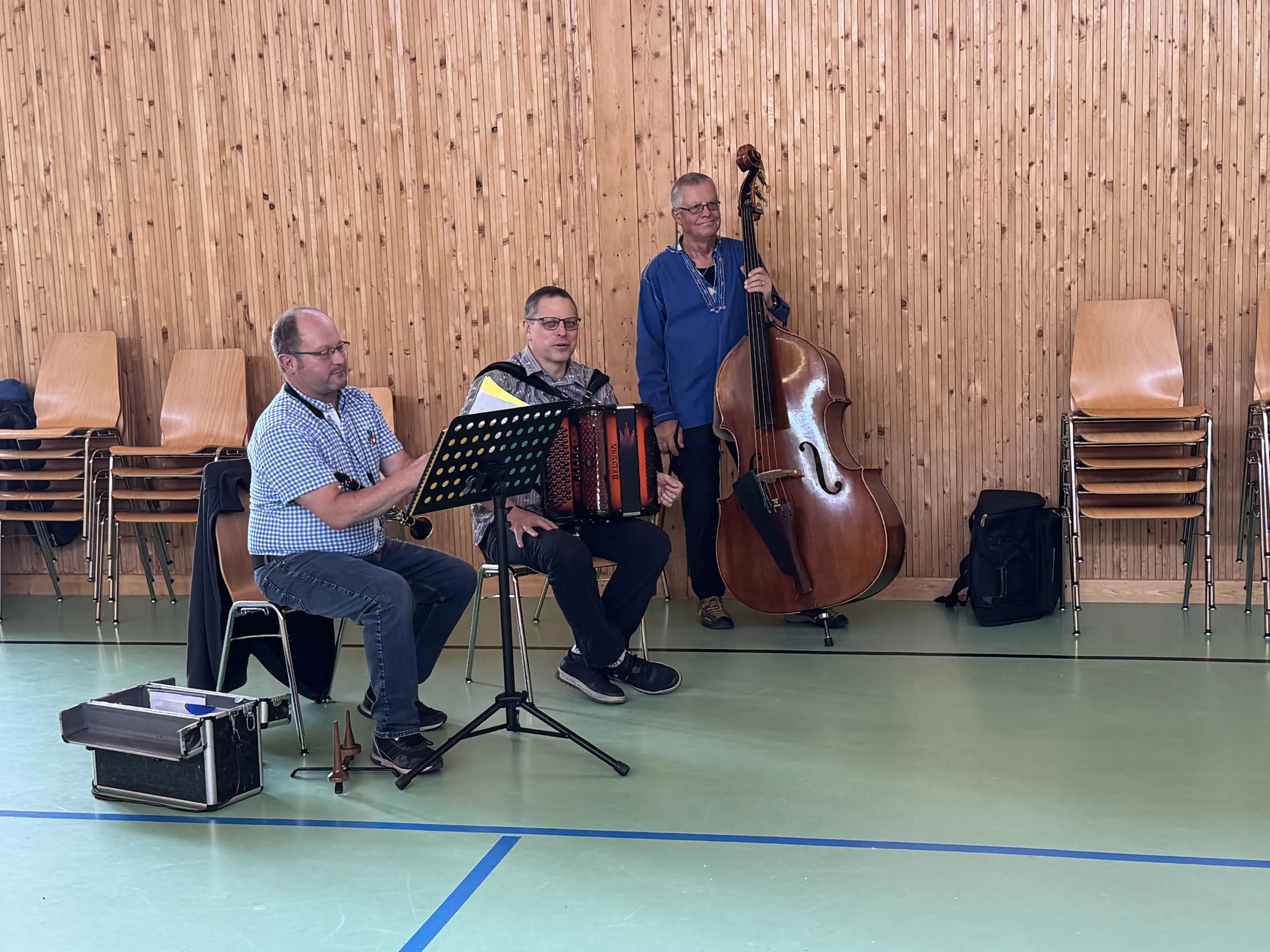 Tre musicisti si esibiscono in una sala con pareti di legno. Un uomo suona l'accordion, un altro suona il contrabbasso, mentre il terzo tiene le note in un leggio. Ci sono sedie impilate sullo sfondo.