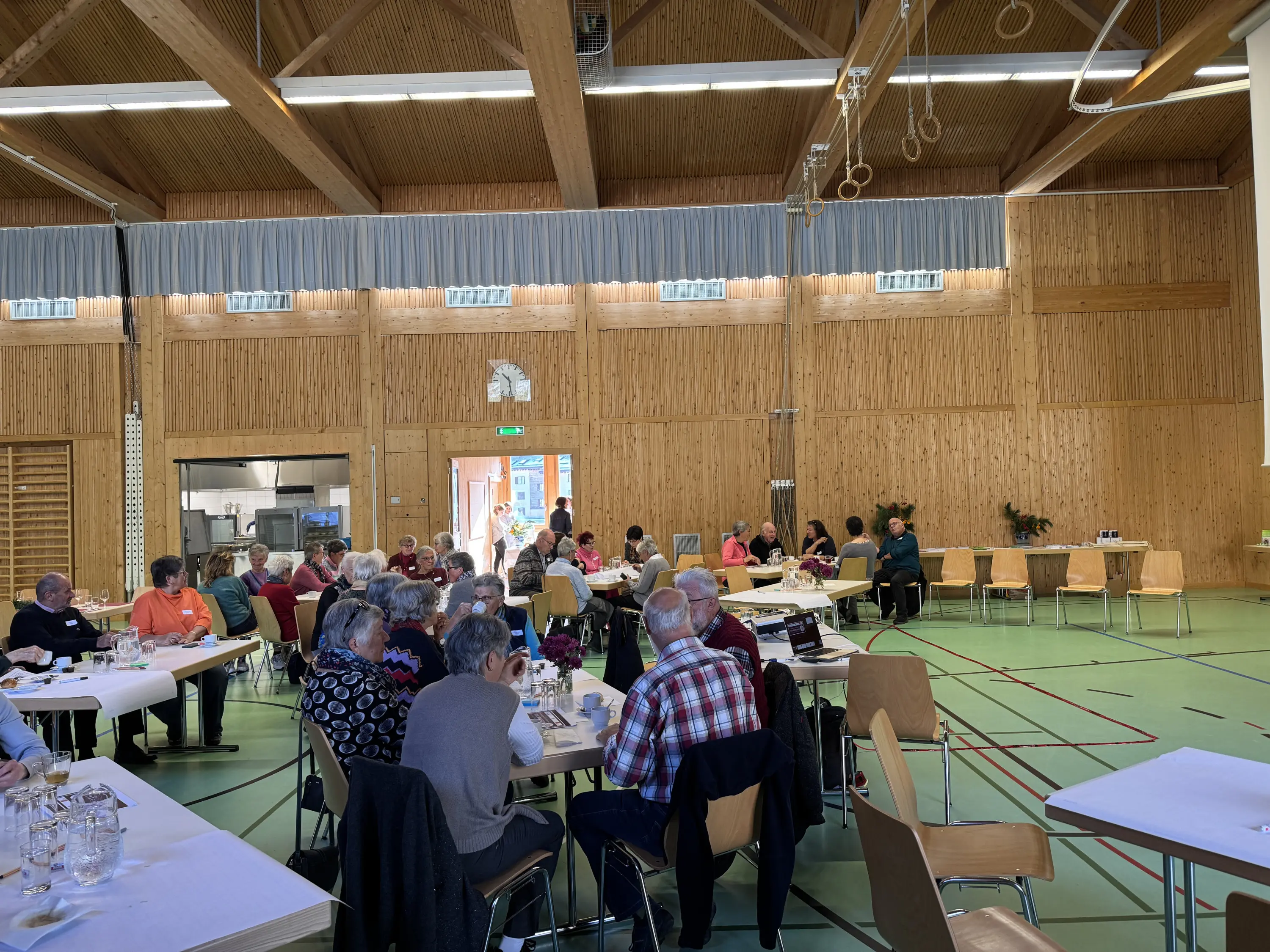 Sala di incontro con molte persone sedute a tavoli, alcune intente a parlare e altre a seguire una presentazione. Ambientazione luminosa e legno a vista.