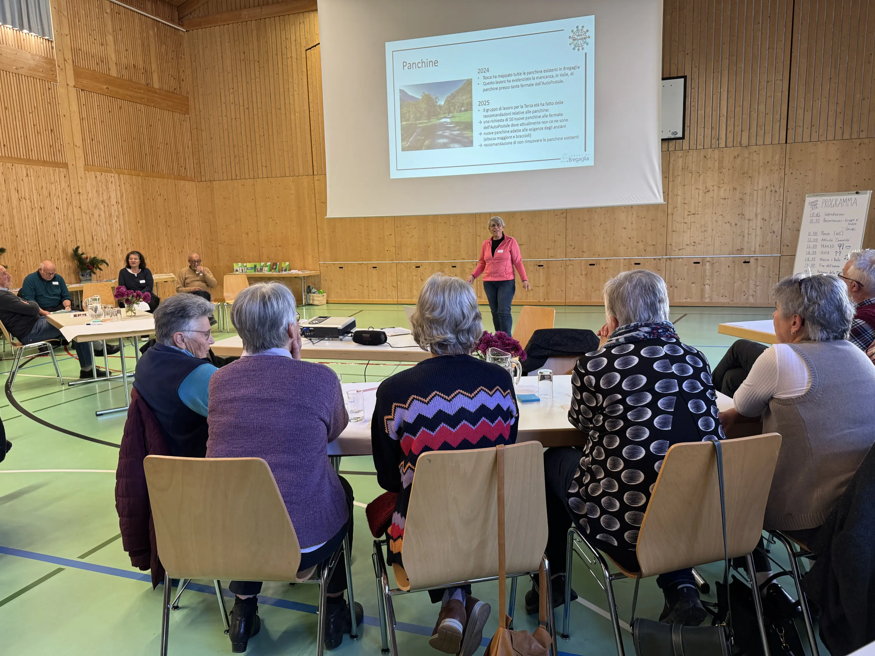 Una presentazione in corso in una sala con diverse persone sedute attorno a un tavolo, mentre una relatrice parla davanti a uno schermo con del testo.