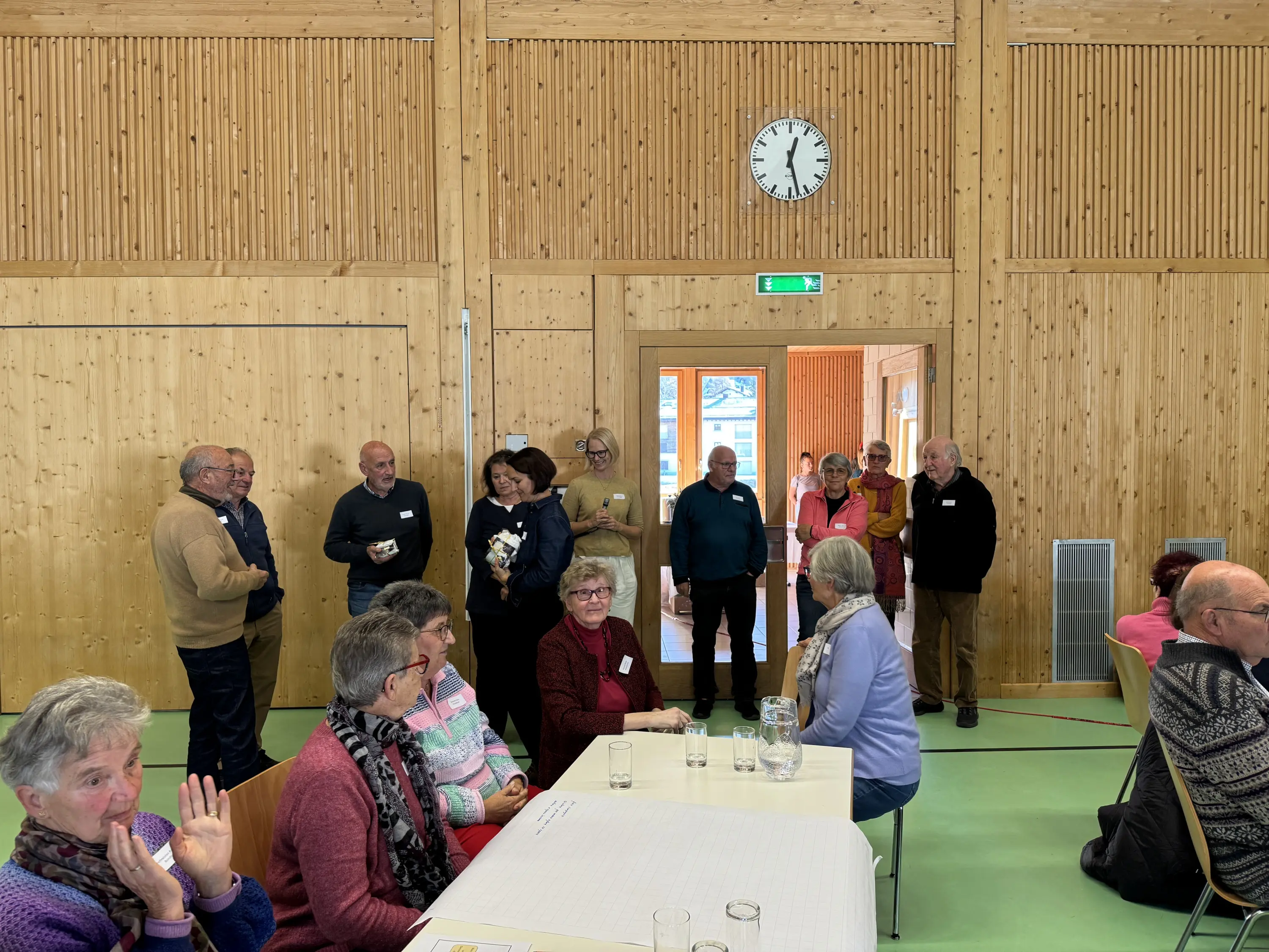 Un gruppo di persone in una sala con pareti di legno si sta interagendo mentre alcuni parlano e altri ascoltano. Sul tavolo ci sono bicchieri di acqua. Un orologio è appeso a una parete.