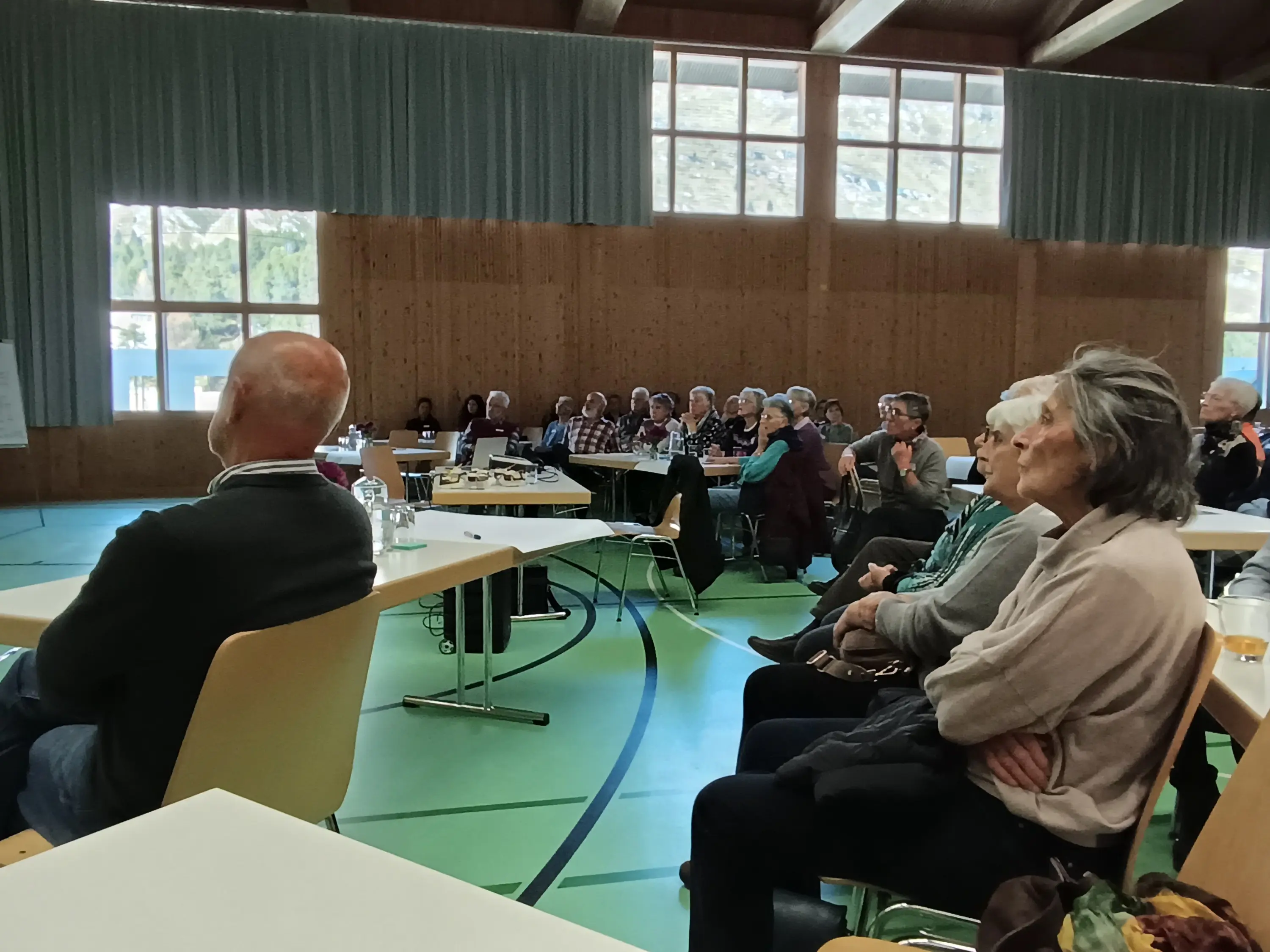 Un gruppo di persone anziane sedute in una sala di conferenze, ascoltando attentamente un relatore. Sullo sfondo, sono visibili schermi e attrezzature audio. La luce naturale entra attraverso le finestre, creando un ambiente accogliente.
