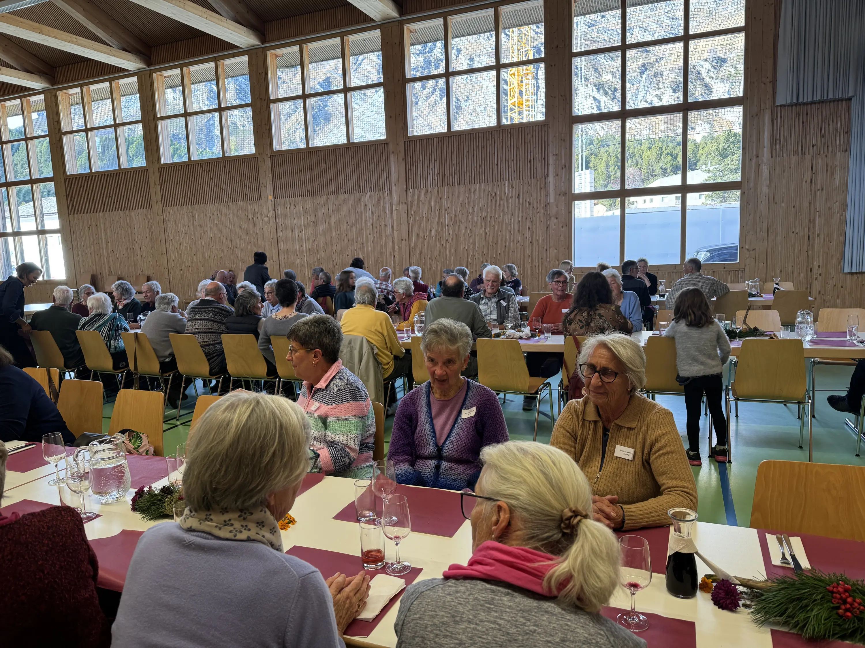 Un grande gruppo di persone sedute a tavoli all’interno di un ambiente luminoso e accogliente. Alcuni partecipanti conversano alle tavole davanti a decorazioni natalizie. Sullo sfondo si vedono altri ospiti seduti e una grande finestra che lascia entrare la