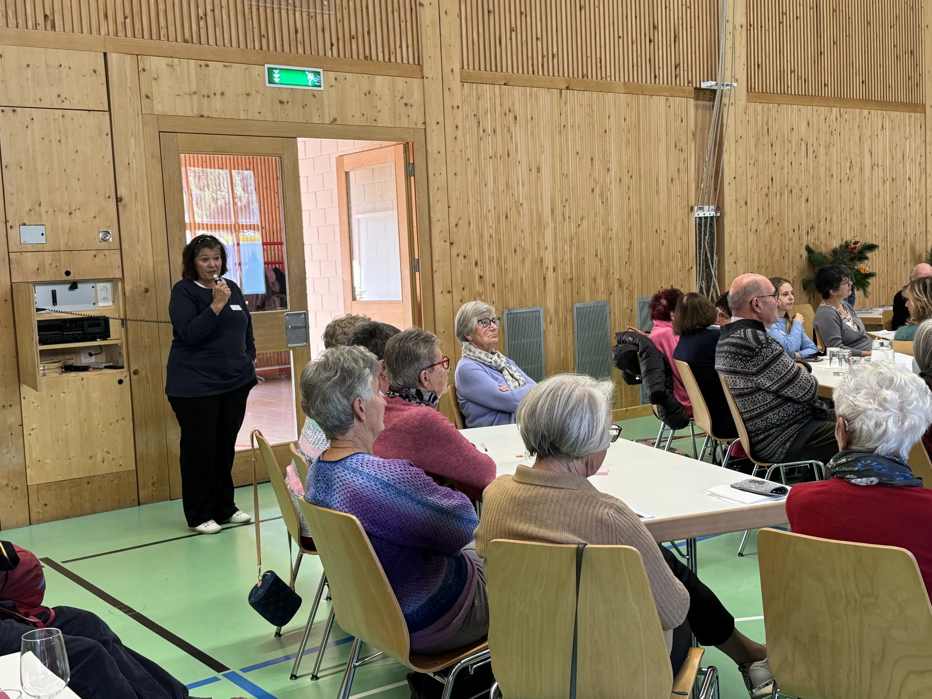 Una riunione in una sala con pareti di legno, dove un'assistente sta parlando a un gruppo di persone sedute attorno a tavoli. Alcuni partecipanti sono di età avanzata e ascoltano attentamente.