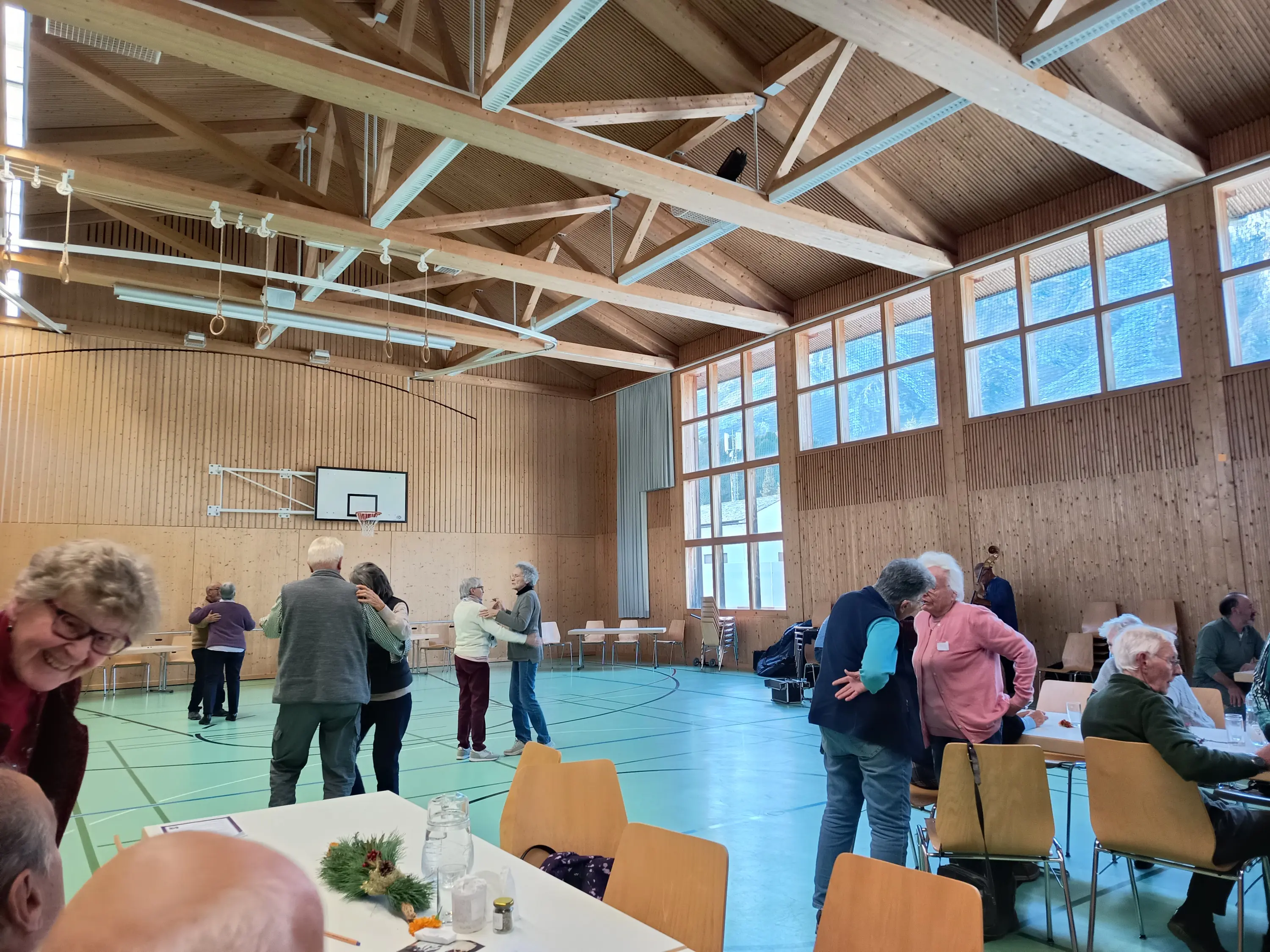 Sala di attività con persone anziane che ballano e socializzano. Le pareti sono in legno chiaro e ci sono grandi finestre che lasciano entrare la luce naturale. Alcune persone sono sedute ai tavoli, mentre altre si muovono nella stanza.