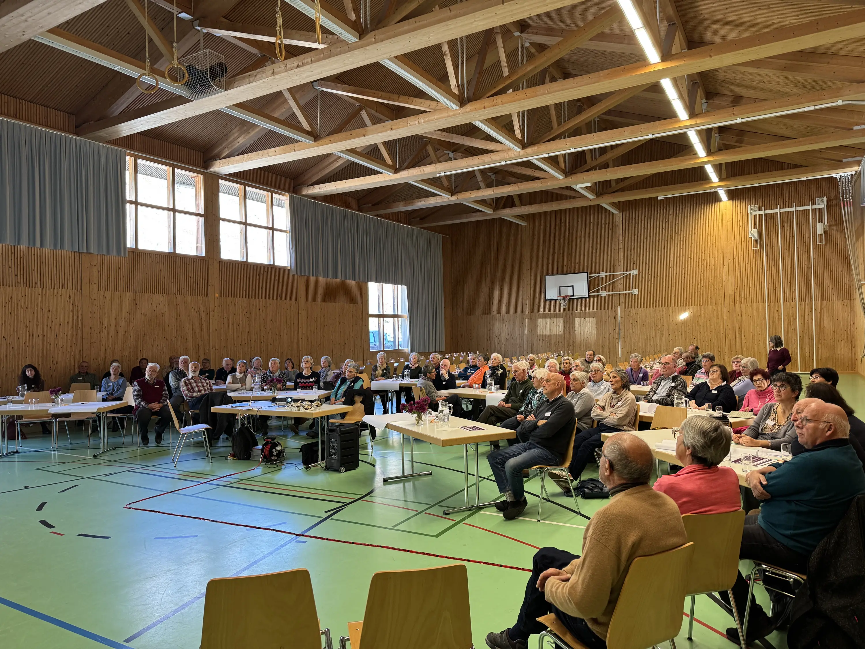 Una sala conferenze affollata con molte persone sedute su sedie, rivolte verso una presentazione. Tavoli con materiali di lavoro sono disposti di fronte ai partecipanti. Il soffitto in legno e le finestre ampie creano un ambiente luminoso.
