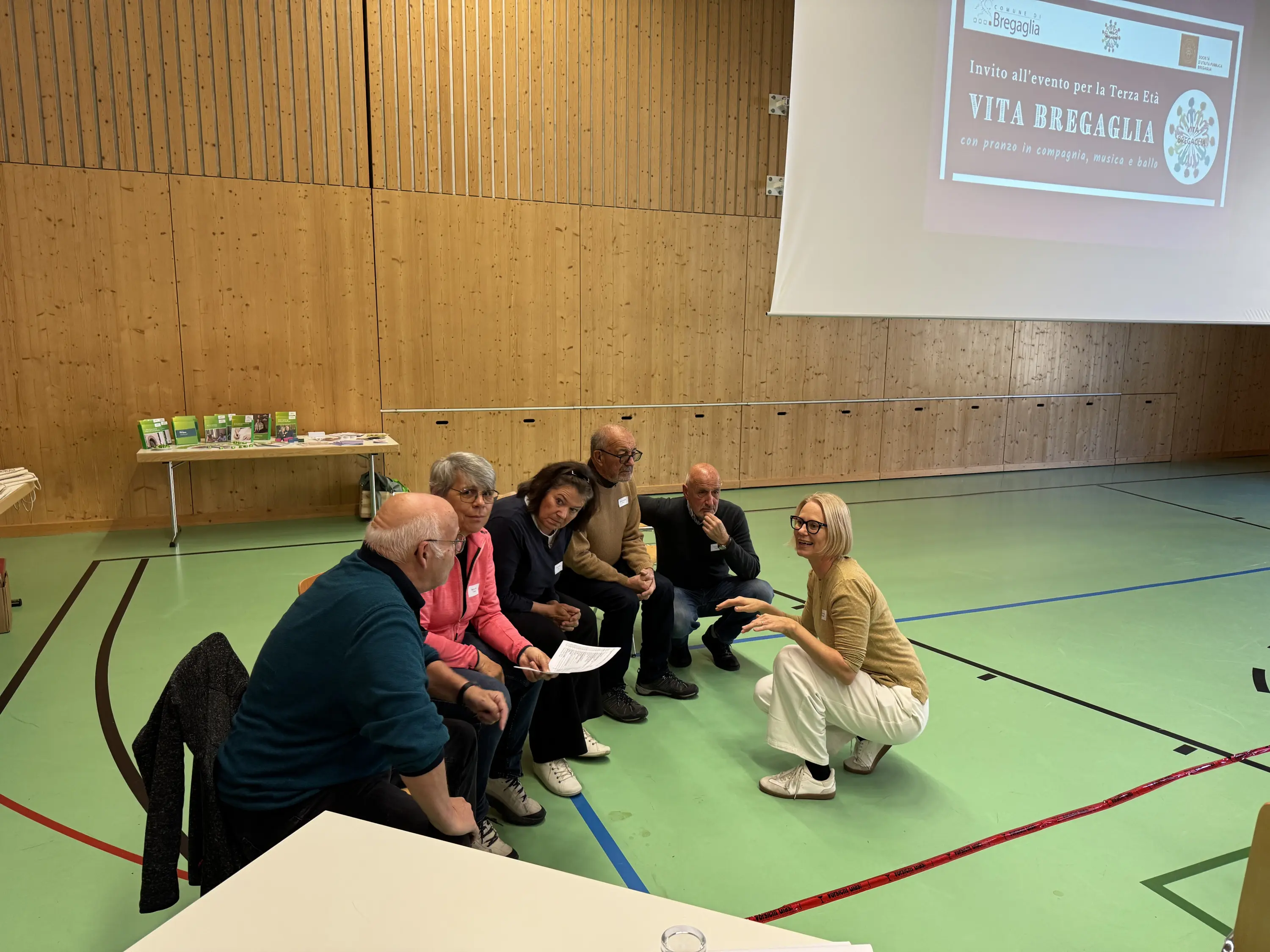 Gruppo di persone sedute attorno a un tavolo in un ambiente interno, discutendo vivacemente. Una donna in piedi sta spiegando qualcosa ai partecipanti. Sullo sfondo, una parete di legno e un proiettore che mostra un banner con il titolo "V
