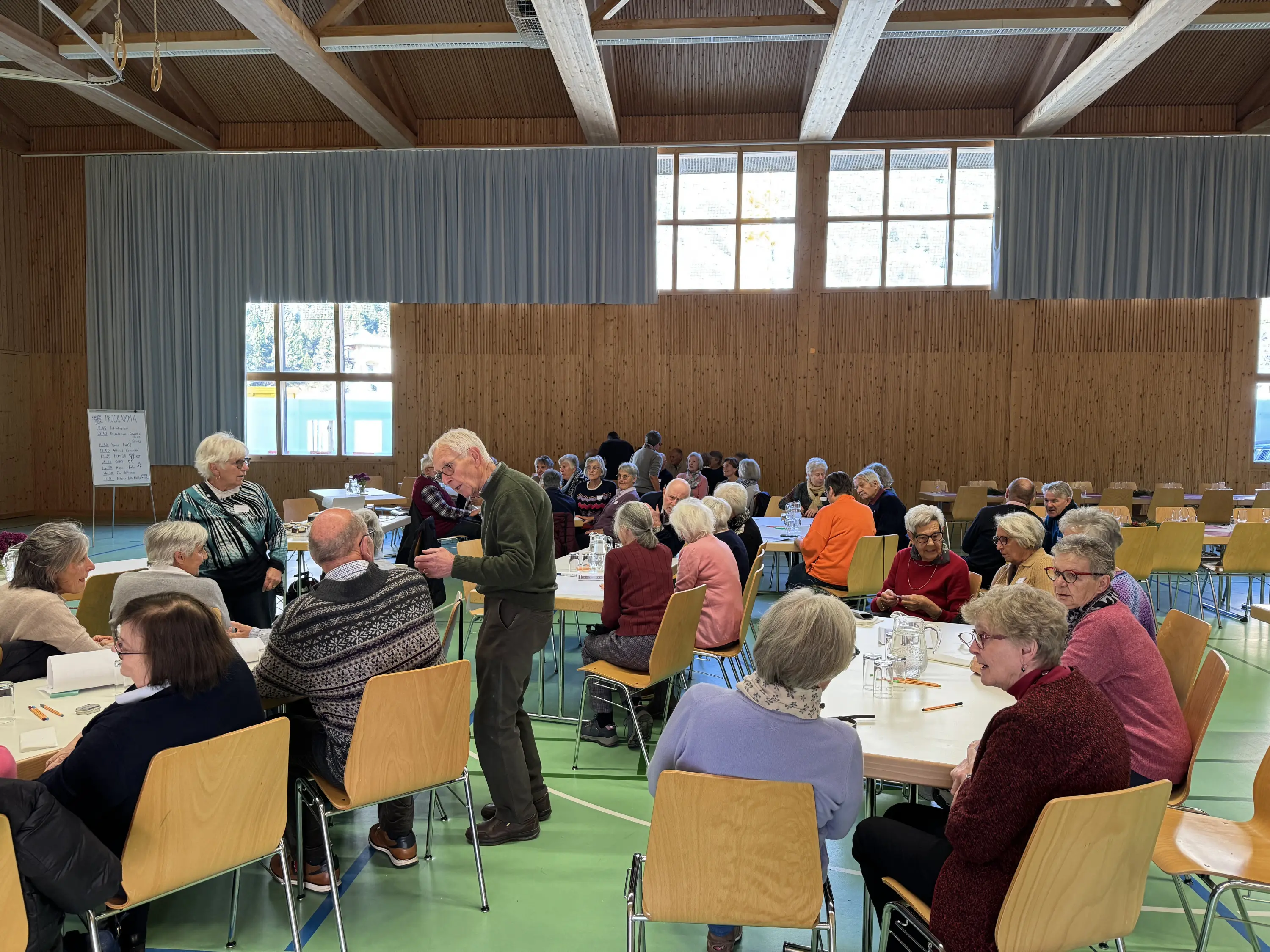 Un gruppo di persone anziane sedute a tavoli in una sala, mentre alcune interagiscono tra loro. La luce naturale entra da grandi finestre e l'ambiente è amichevole e accogliente.