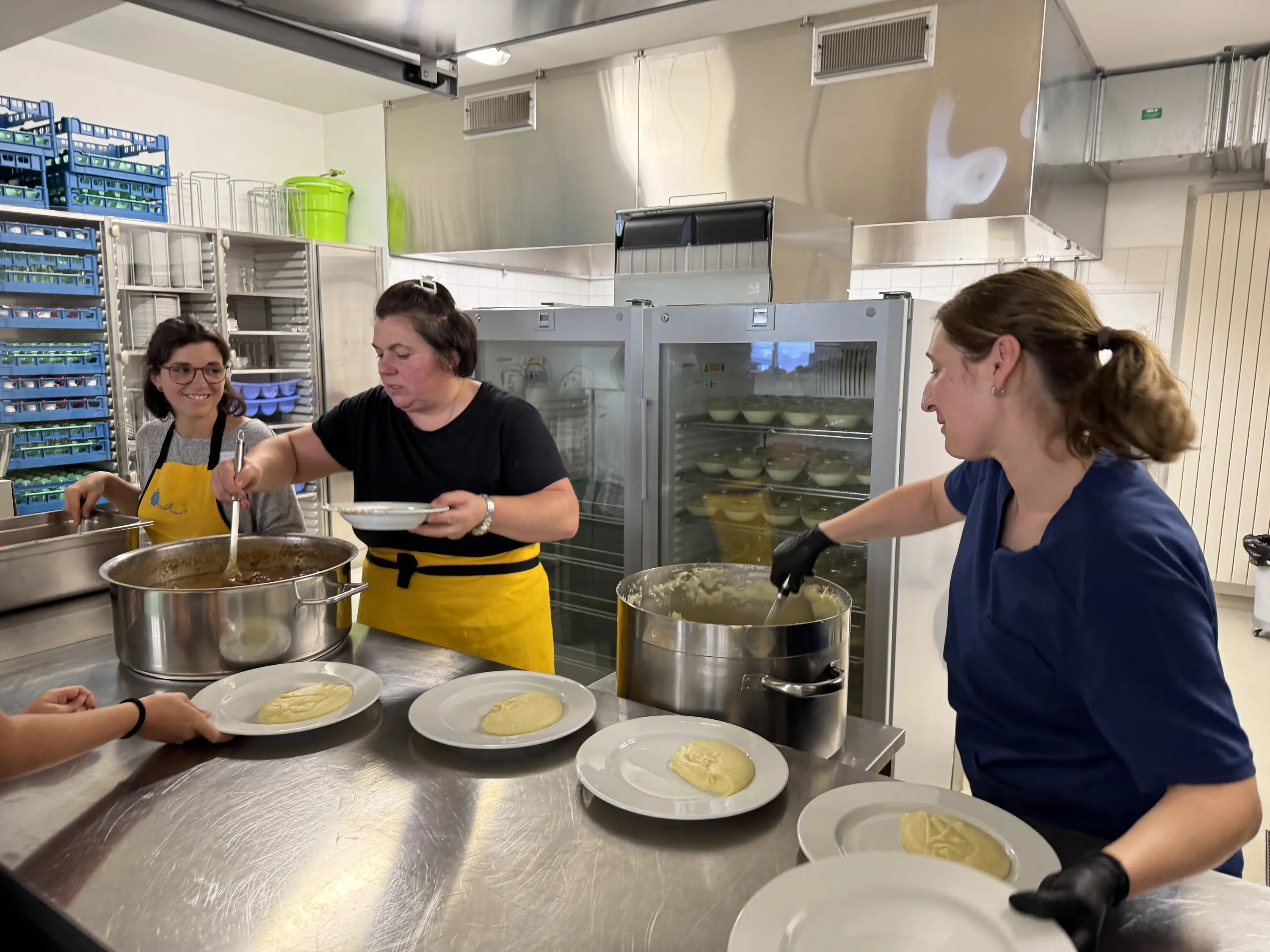 Tre donne in una cucina industriale stanno servendo cibo su piatti bianchi. Una donna versa un sugo in un piatto, mentre un'altra prende una porzione di purè di patate, e la terza osserva sorridendo. Sullo sfondo si vedono