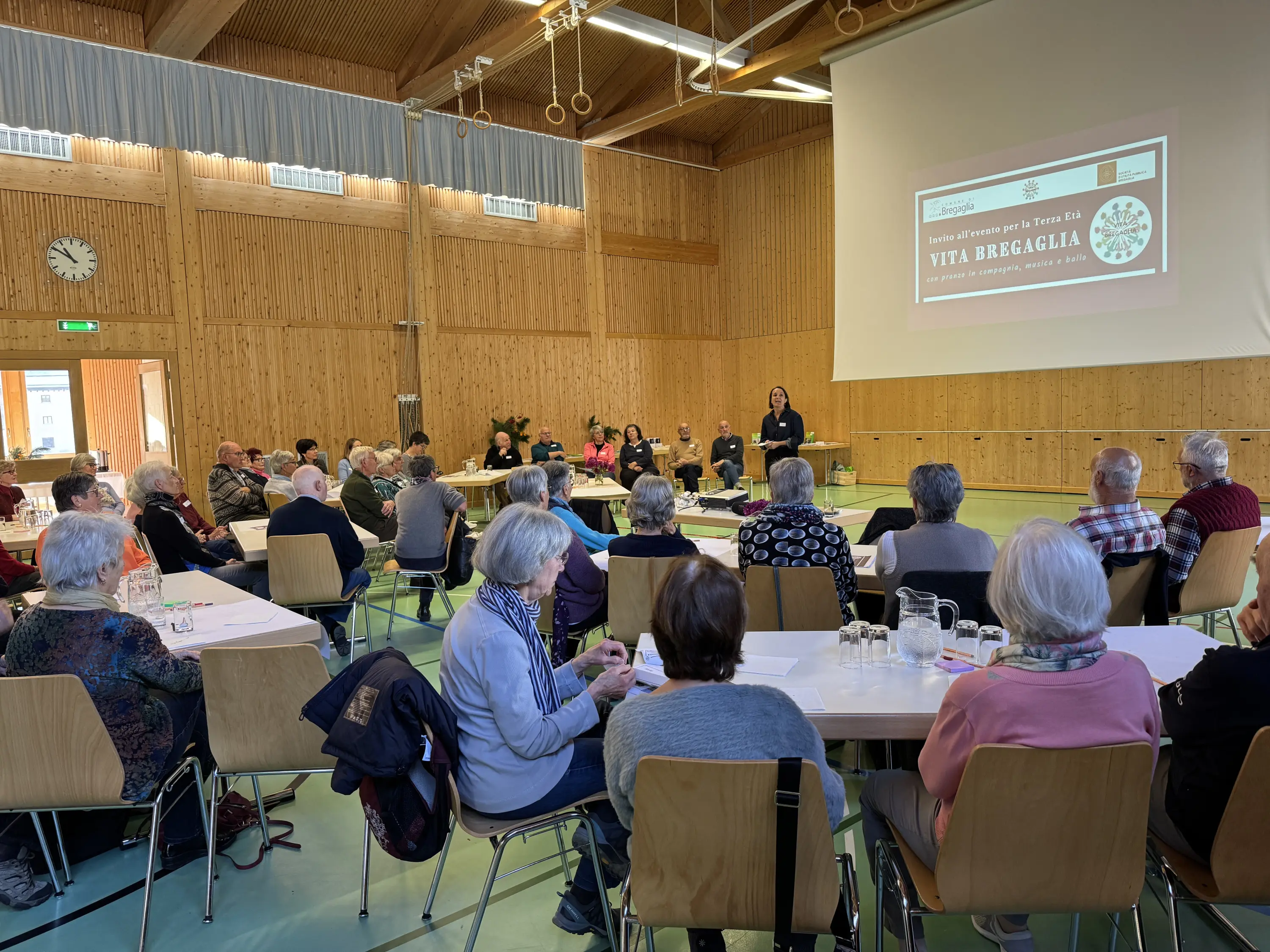 Un gruppo di persone anziane sedute in una sala riunioni, mentre ascoltano un relatore in piedi davanti a un proiettore. Sullo sfondo, un grande schermo mostra un titolo e un programma. L’atmosfera è conviviale e coinvolgente.